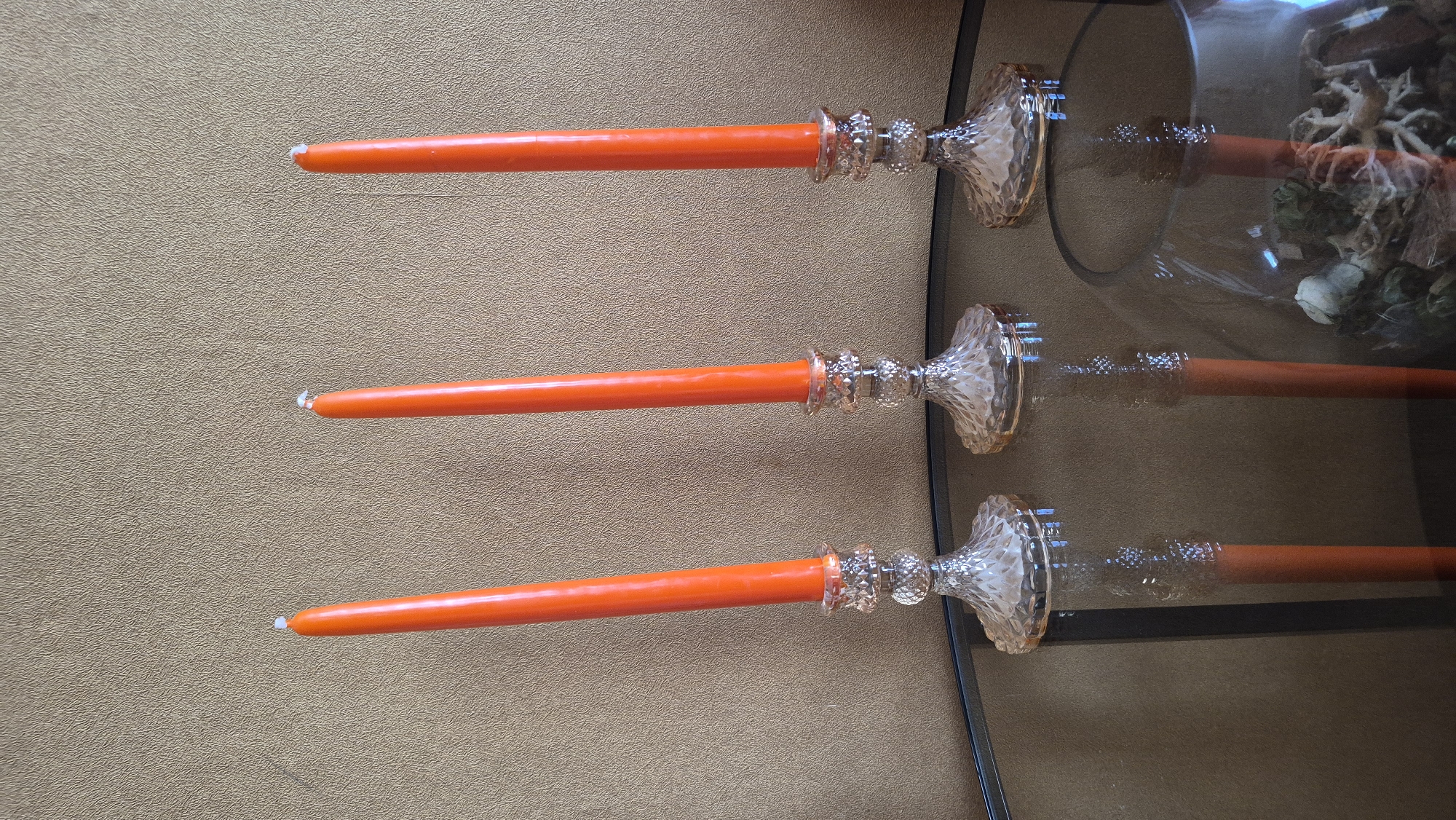 Three orange taper candles stand in ornate glass candle holders on a dark glass tabletop. The candle holders have faceted bases and stems. The background is a textured, brown wall. The scene is indoors, likely in a home. The location of Krauja, Latvia is not discernible from the image.