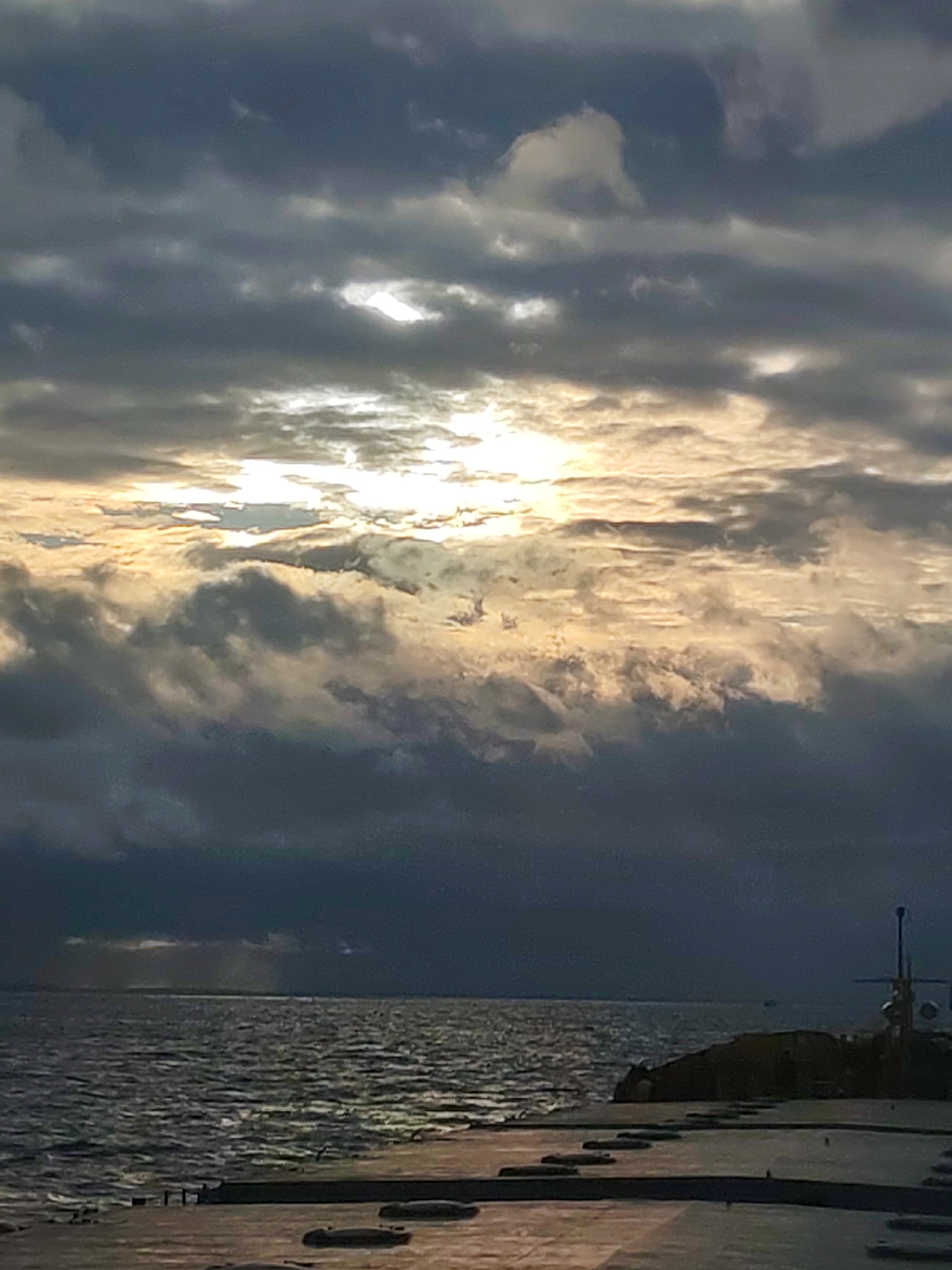 The image shows a seascape under a cloudy sky, with the sun partially obscured. The water is dark with visible ripples and waves. A concrete structure is in the foreground, likely a pier or seawall. There is a structure with antennas extending upwards towards the right side of the image. The location is unknown.