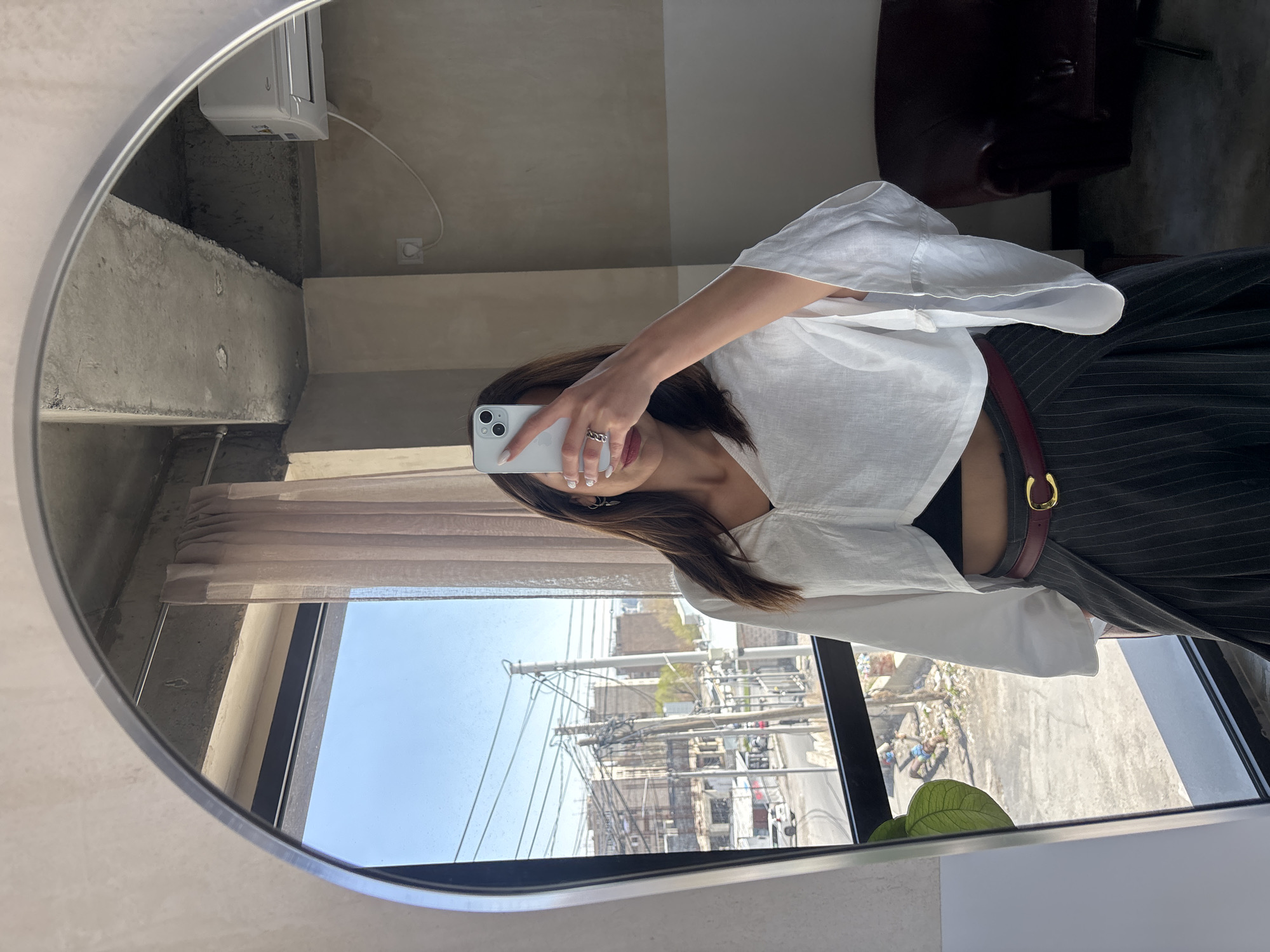 A mirrored selfie shows a woman wearing a white blouse and striped pants with a red belt. She holds a white smartphone, partially obscuring her face. The reflection includes the interior of a room with concrete elements and a window revealing a cityscape. The urban landscape includes buildings, power lines, and visible litter. The location is Yerevan, Armenia.