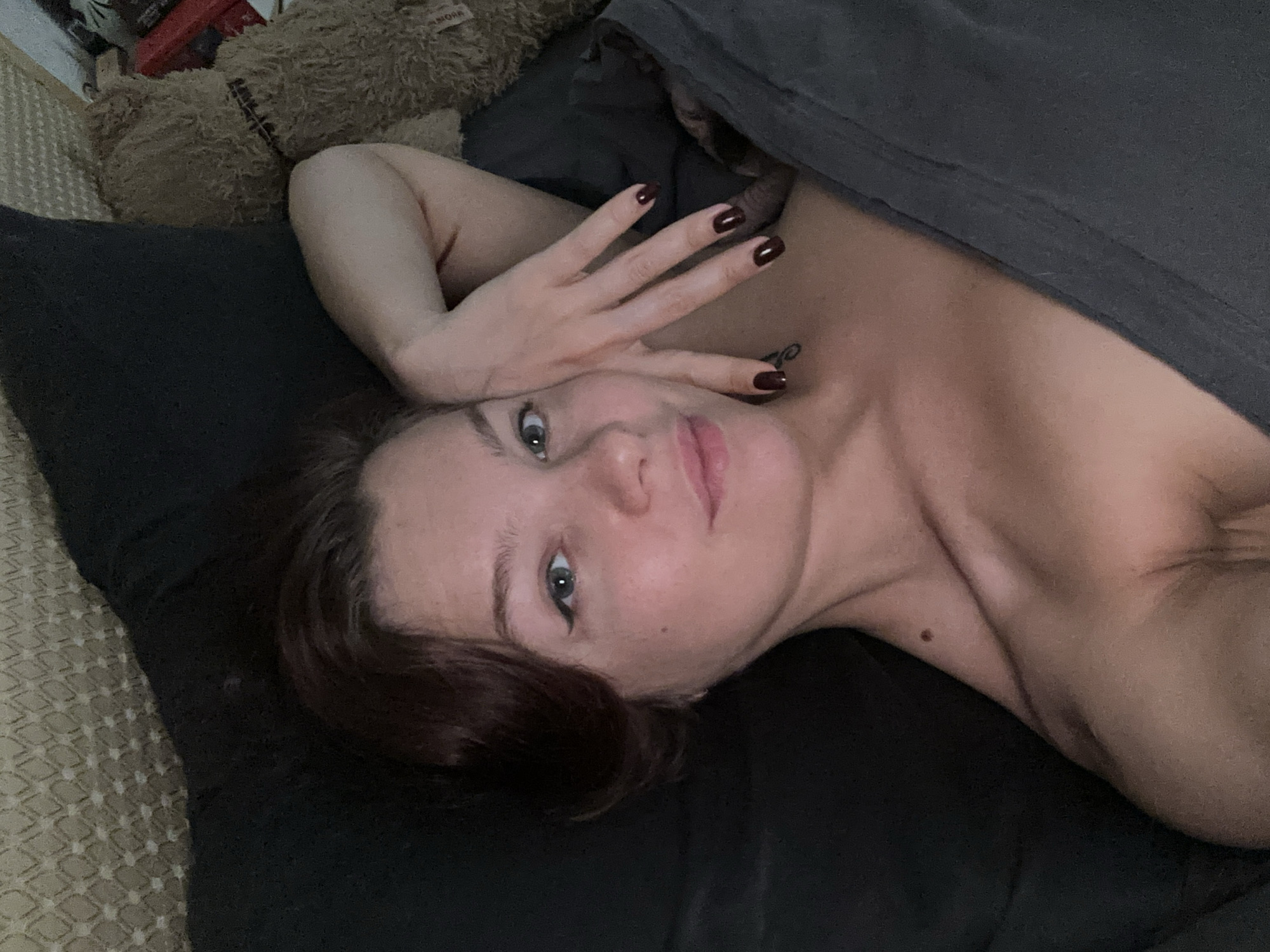 A woman is lying on a bed, partially covered by a dark gray sheet. Her head rests on a dark pillow. She has short, dark hair and fair skin. Her left hand is raised and positioned near her forehead, showing dark polished fingernails. There is a tattoo visible on her upper chest. A brown teddy bear is located in the upper left corner of the frame. The location cannot be determined from the image.