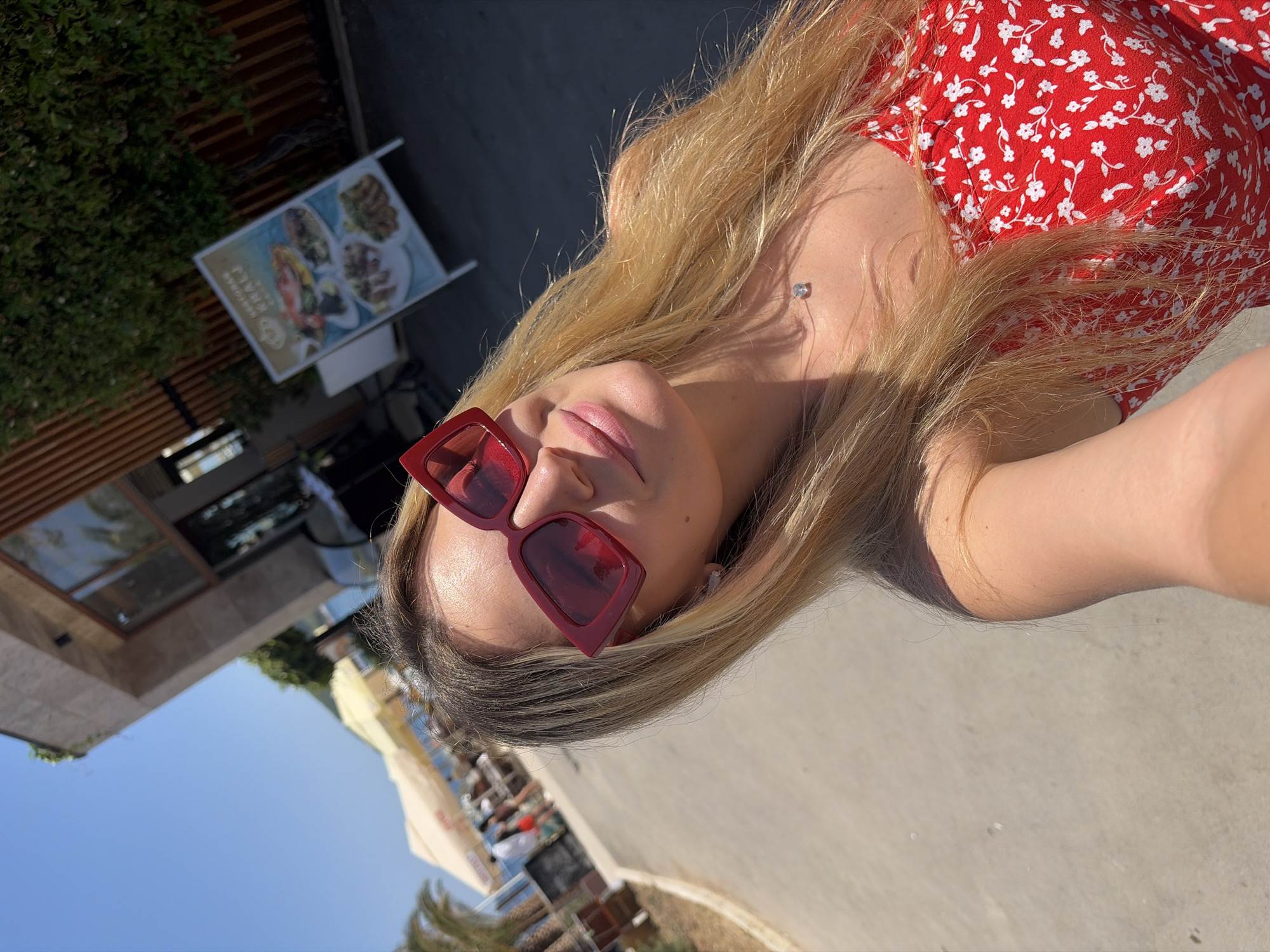 An outdoor, sunny, eye-level selfie is taken of a blonde woman with fair skin, wearing large, angular red-framed sunglasses and a red floral dress. She is looking directly at the camera with her head tilted slightly to her left. Her hair is long and blonde, framing her face. A delicate, clear pendant necklace is visible around her neck. The background shows a building with stone and wood accents, a bright blue sky, and palm trees. To the right of the building, a freestanding sign displays images of various dishes. In the distance, there are beach umbrellas and people. The ground is paved. The overall scene suggests a warm, vacation-like atmosphere, likely in a coastal city like Budva, Montenegro, given the presence of beach umbrellas and the Mediterranean-style architecture.