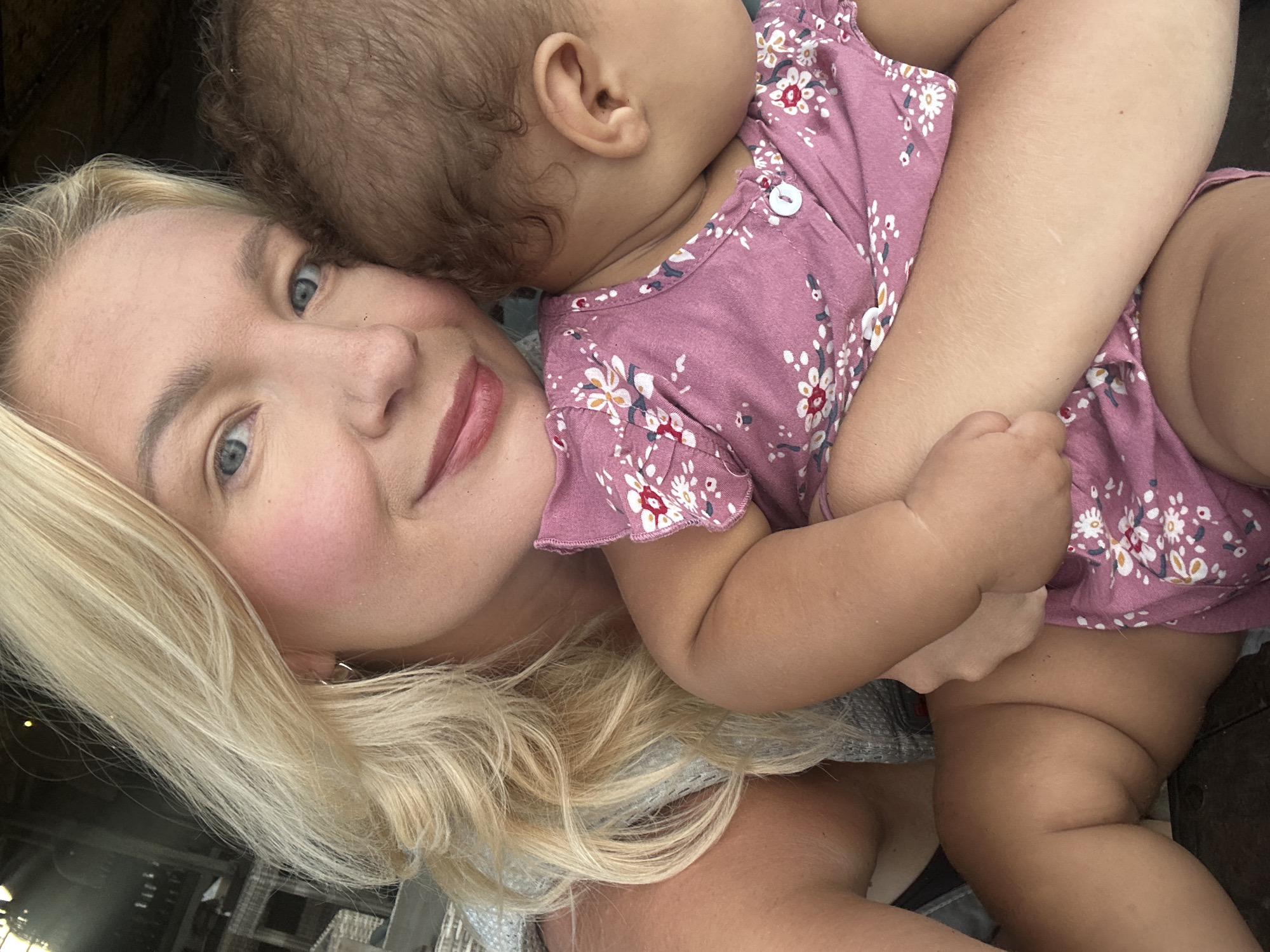 A Caucasian woman with blonde hair and blue eyes is holding a baby with brown hair. The woman is wearing a gray top and the baby is wearing a pink floral dress. They appear to be indoors. The background is out of focus but suggests an indoor environment.