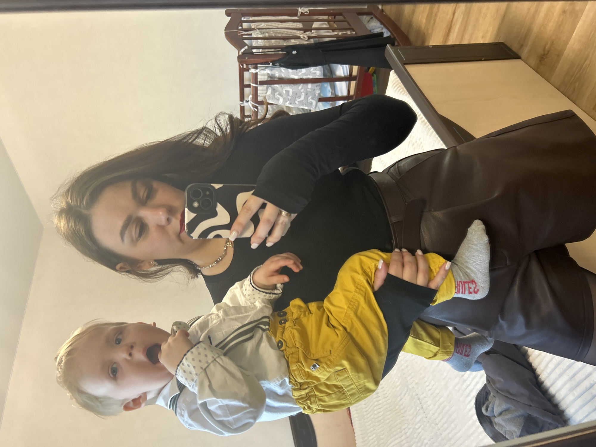 A woman is taking a selfie in a mirror while holding a baby. The woman has long brown hair and is wearing a black long-sleeved shirt, a silver necklace, and brown leather pants. She is holding a smartphone with a zebra-striped case. The baby has blonde hair and is wearing a white and gray striped long-sleeved shirt and yellow pants. They are both looking at the camera. The background includes a crib and a bed. The scene appears to be indoors. There is no information to confirm this image was taken in Chaikovsky, Russia.