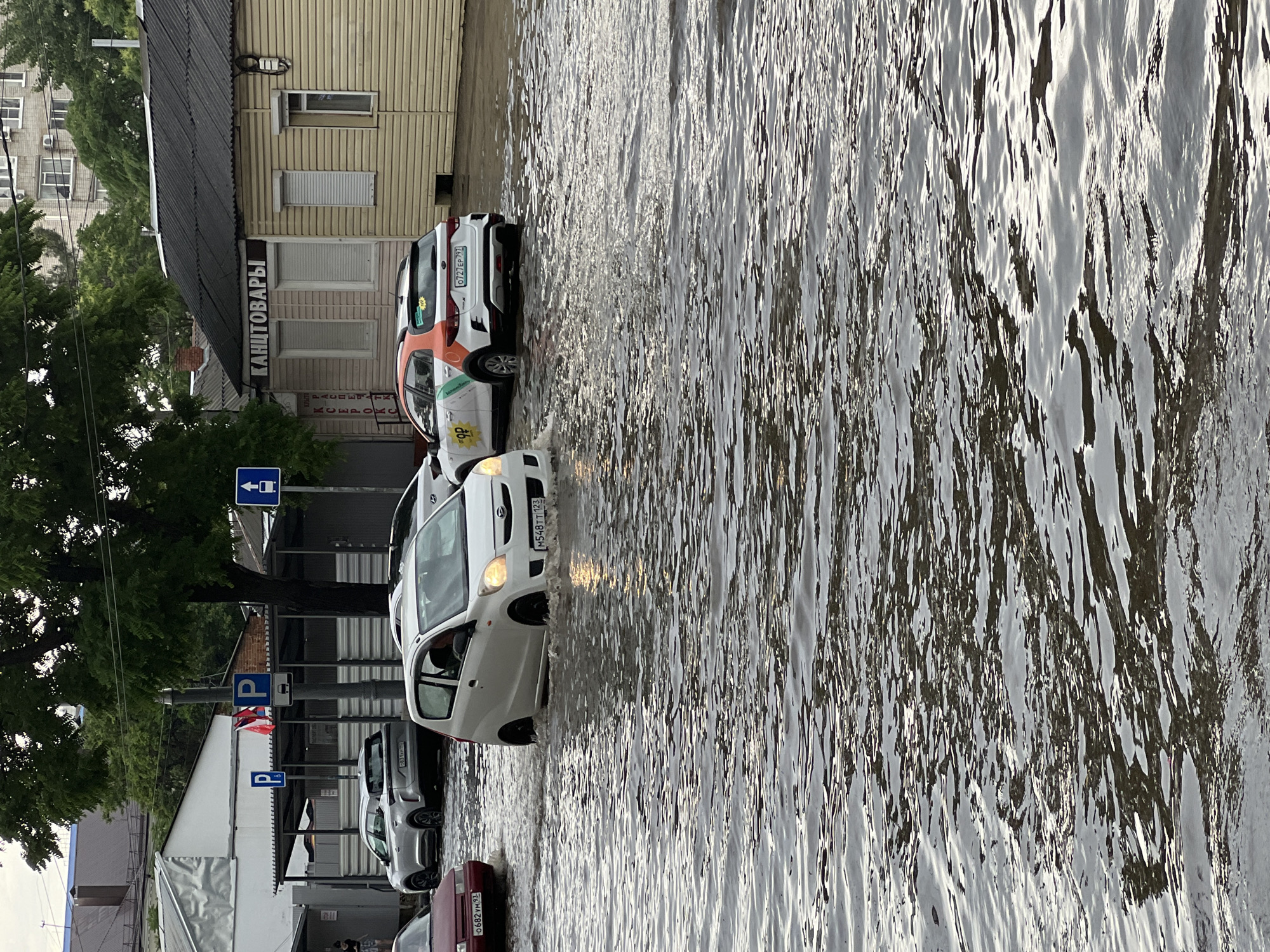 On a street in Krasnodar, Russia, several cars are driving through a flooded street. The water level is high enough to submerge the lower parts of the vehicles. One white car has its headlights on and is creating a wake as it moves through the water. Another car, which appears to be a taxi with orange and green markings, is also visible in the flooded street. The surrounding buildings suggest an urban environment, with one building displaying a sign that reads "КАНЦТОВАРЫ" (Kantsotvary), which translates to "Stationery." There are trees and street signs visible, indicating this is an outdoor scene. The overall impression is one of heavy rainfall and its impact on the urban infrastructure.