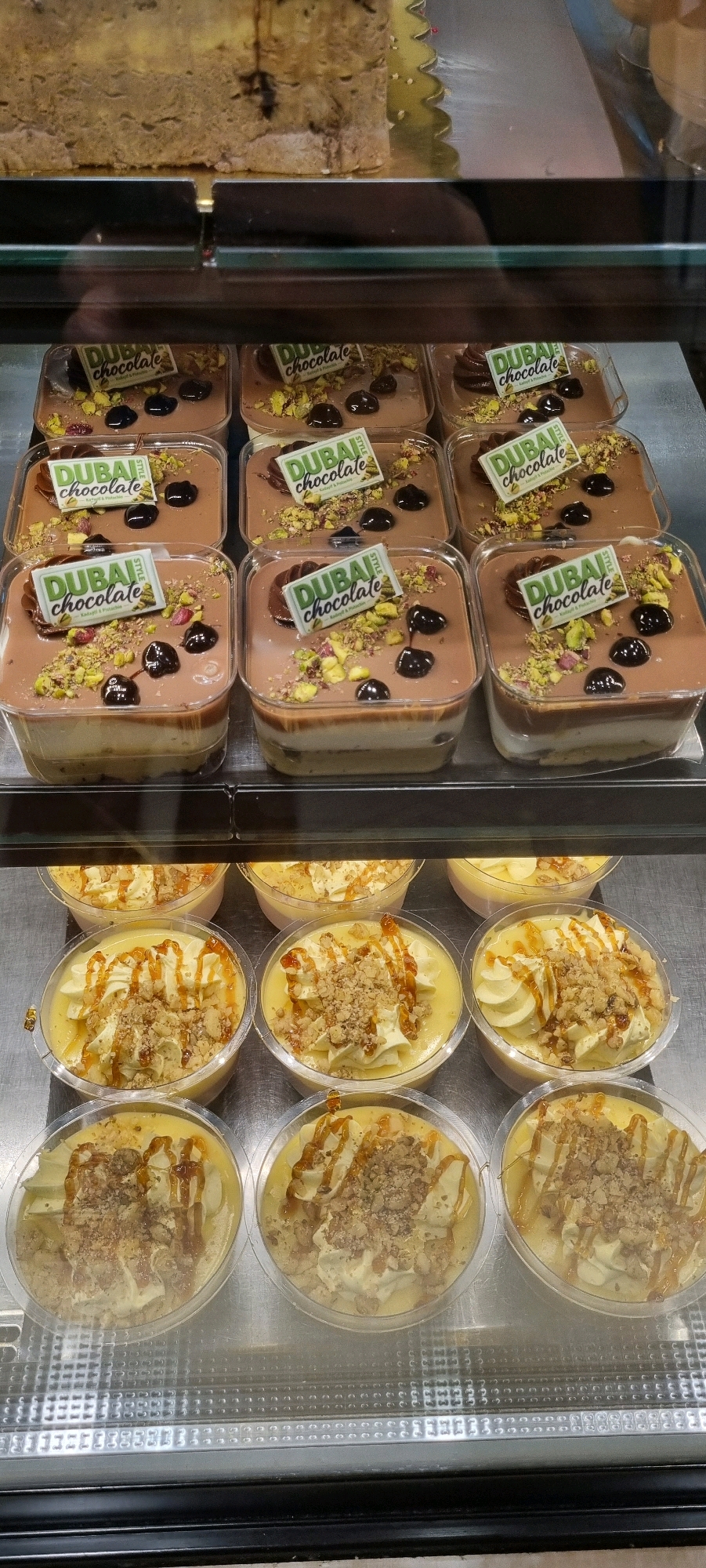 The image shows a display of desserts in plastic containers inside a glass-enclosed refrigerated case. The top shelf features a large, multi-layered dessert, possibly a cake or loaf, with visible crumbs and what appears to be chocolate or dark fruit within its layers. Below this, the middle shelf displays numerous individual dessert portions. These desserts are layered, with a light-colored base and a darker, smooth topping. Each of these portions is adorned with chopped pistachios, dark chocolate dollops, and a small, green and white "DUBAI STYLE CHOCOLATE" wrapper. The bottom shelf holds more individual desserts in clear plastic cups. These appear to be a creamy, yellow-hued dessert topped with whipped cream, drizzled caramel sauce, and sprinkled with crunchy bits, possibly nuts or cookie crumbles. The overall scene suggests a confectionery or bakery display in Bashkia Durrës, Albania, where these items are arranged for sale. There are no people or animals visible in the image.