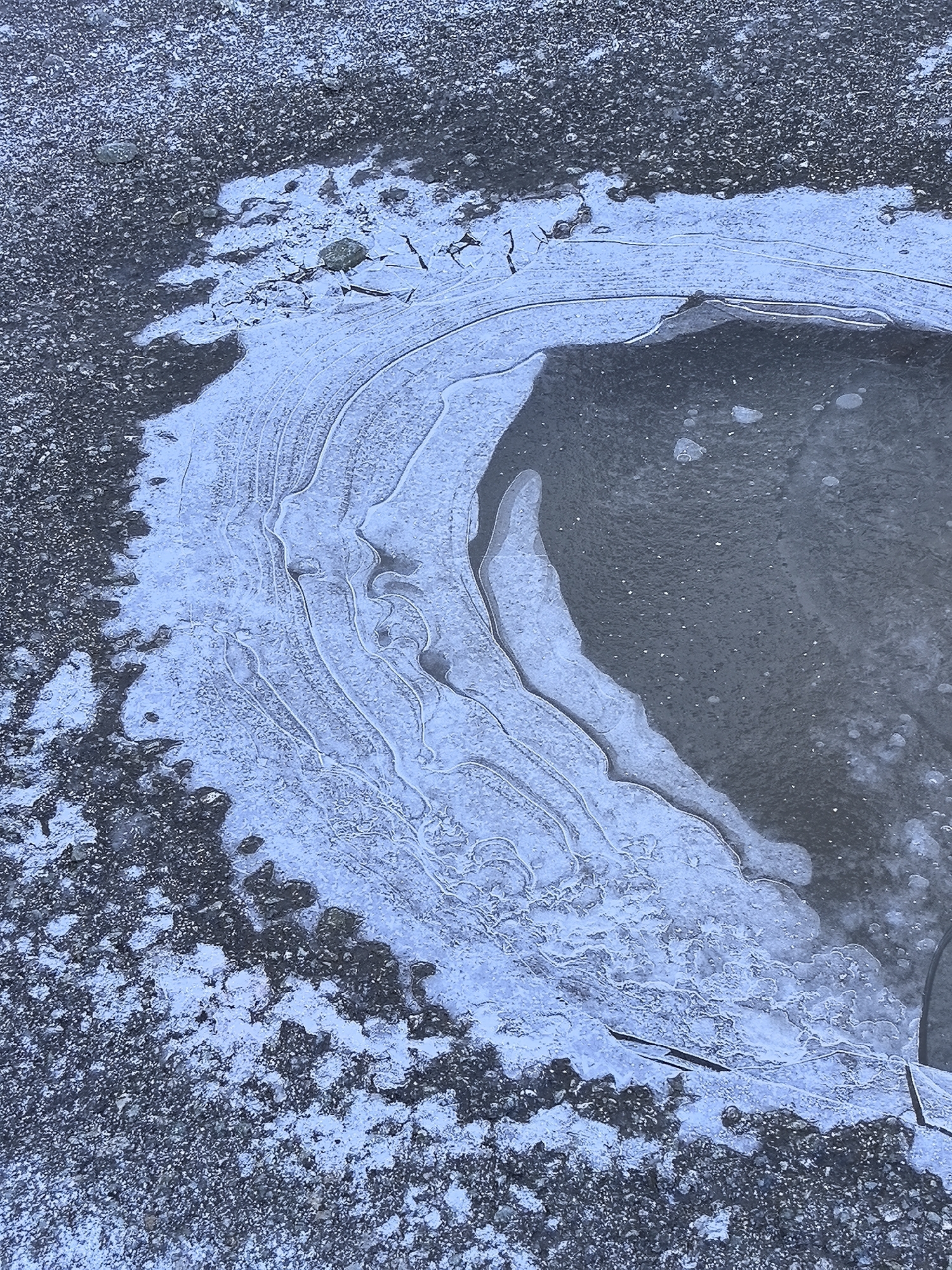 A high-angle close-up photograph depicts a patch of ice and snow-covered ground. The ice covers the ground, forming a semi-circular shape with layered patterns. The ice is translucent in some areas, revealing the ground underneath. The ground surrounding the ice patch appears rough and is covered with a mix of snow and dirt. The location in the city is unknown.