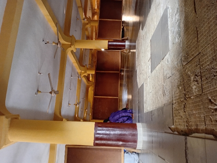 The interior showcases rows of wooden pews under a high ceiling supported by yellow-painted beams. Several ceiling fans are evenly spaced along the ceiling. The floor features a mix of smooth tiles and a rougher, potentially unfinished section near the wall. A purple jacket lies on one of the pews. The perspective gives a somewhat distorted view, emphasizing the height of the room and the repetition of the architectural elements.