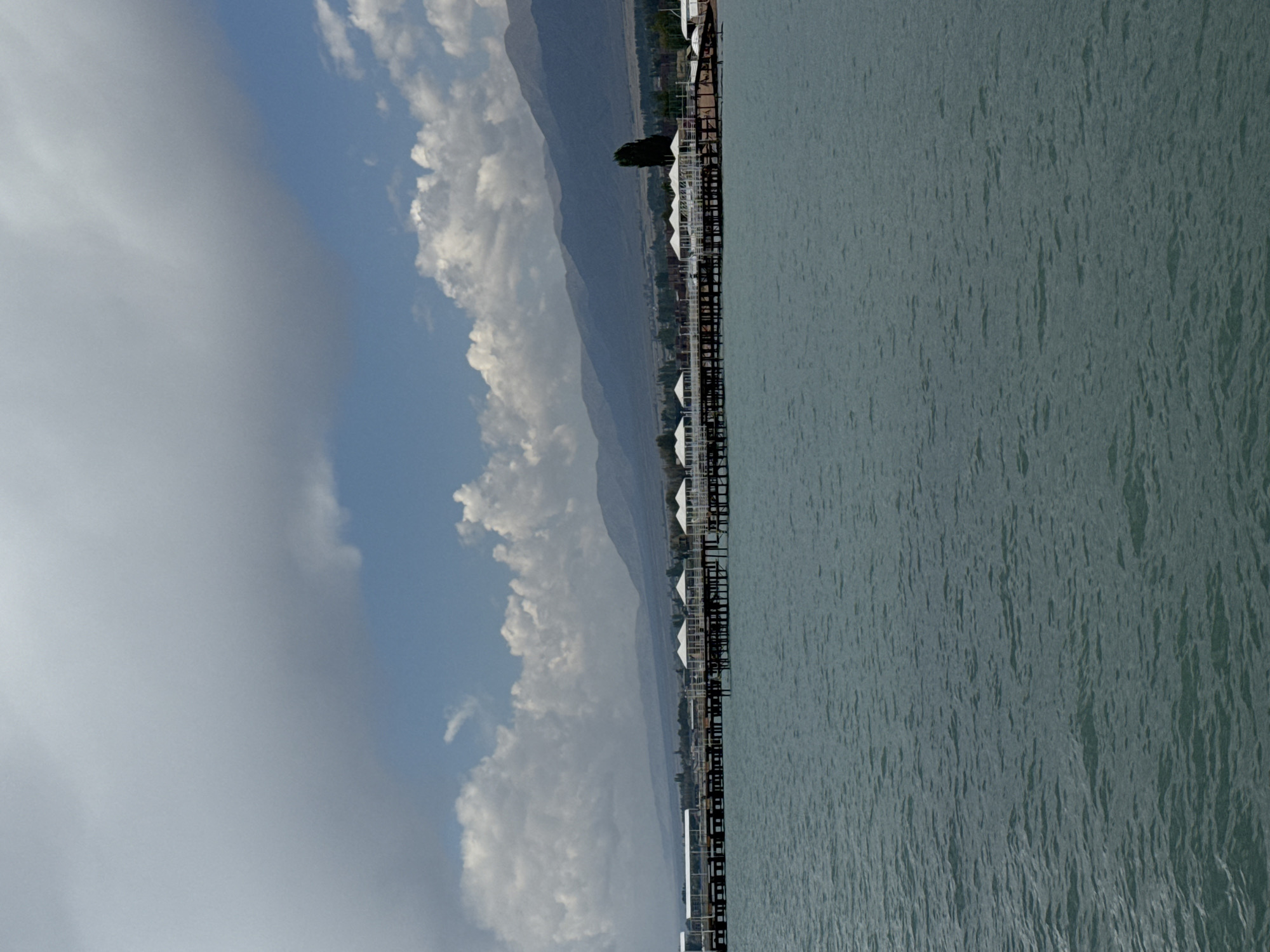 The image depicts a serene lake scene in Sary-Oy, Kyrgyzstan, with a long pier extending into the water. The pier is lined with several white, open-sided gazebos, offering shelter and a picturesque view of the lake. In the background, a range of mountains stretches across the horizon, their slopes softened by distance and a hazy atmosphere. The sky above is a mix of blue and white clouds, with larger, darker clouds dominating the upper portion of the frame. The water is a deep blue-green, with gentle ripples indicating a calm day. The overall impression is one of tranquility and natural beauty.