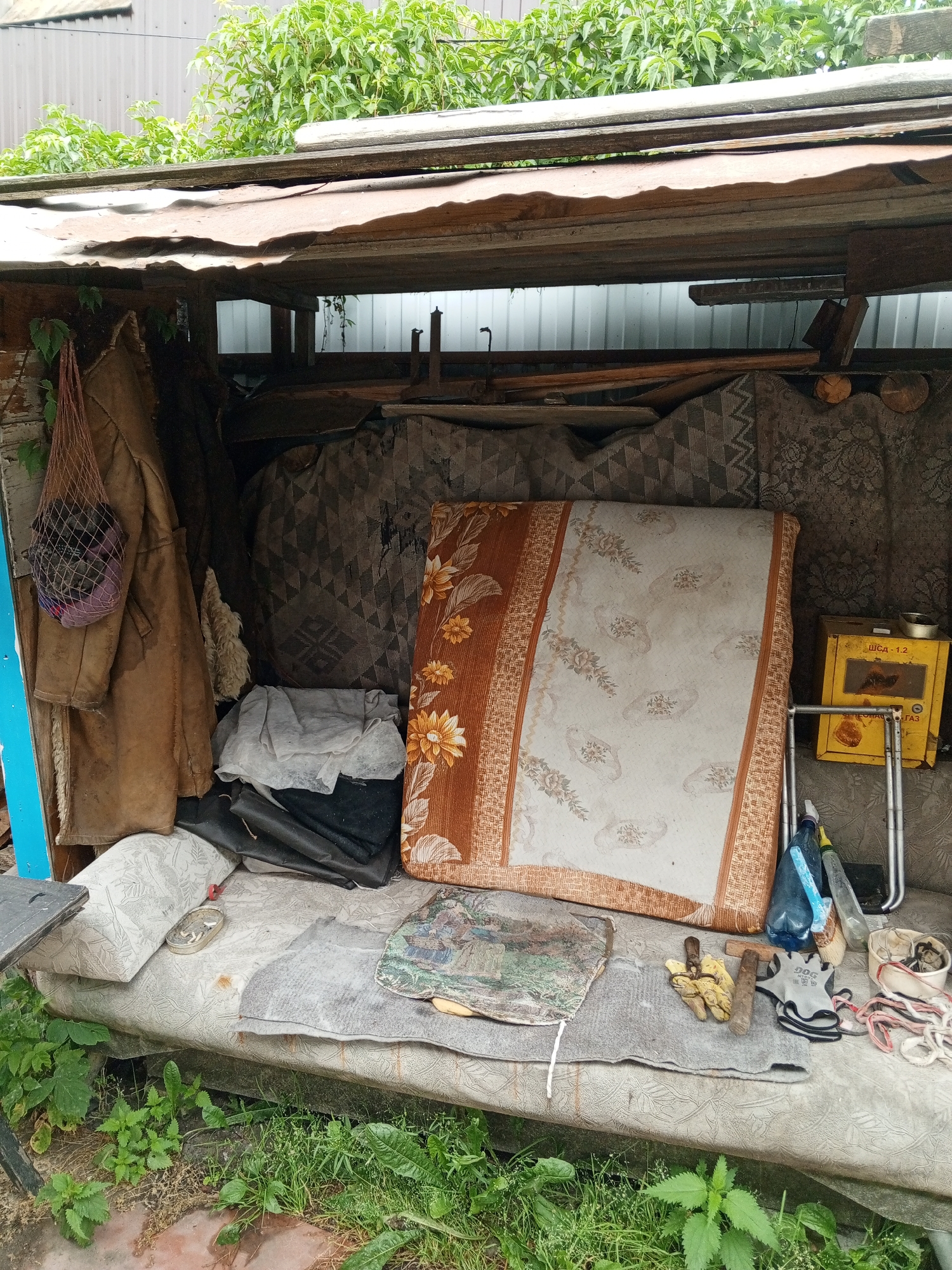 The image depicts an outdoor storage area, possibly a makeshift shed or lean-to, filled with various discarded items. In the foreground, a worn-out mattress serves as a base for a collection of objects. On top of the mattress, a grey rug is laid out, with a small framed picture resting on it. Adjacent to this, a pair of yellow work gloves and a pair of black and grey work gloves lie next to a wooden handle. To the right, a yellow metal box with Cyrillic lettering is visible, along with some plastic bottles and a white bucket containing string.
Behind the mattress, stacked items include what appears to be a folded mattress or blanket with a floral pattern. A brown coat hangs from a hook on the left, with a net bag containing a purple object suspended from it. Several large, patterned fabric pieces are propped up against the back wall, resembling upholstery or blankets. The overall environment is cluttered and suggests a space used for storage of miscellaneous items. The location is identified as Shadrinsk, Russia.