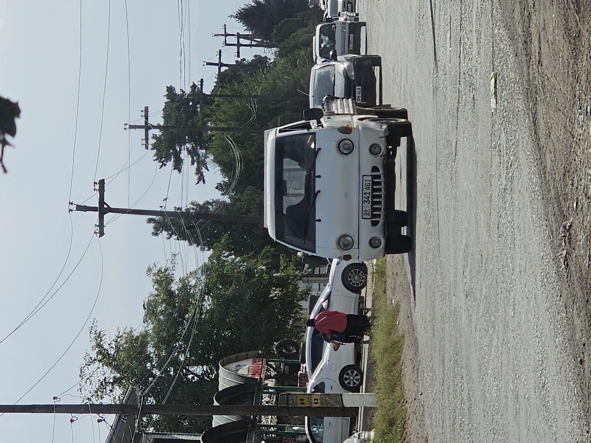 In Özgön, Kyrgyzstan, on a dusty, paved street under a hazy sky, a white, flatbed truck drives down the road. The truck has a license plate that reads "06 341 AG1 KG". Several other vehicles are also visible in the distance, including a white car, a dark sedan, and other trucks. On the left side of the street, there are a few market stalls with arched roofs and some trees. A woman wearing a pink shirt and a dark skirt, with a child standing next to her, is visible near the stalls. Utility poles and power lines crisscross the sky. The overall scene depicts a typical day in a Central Asian town.