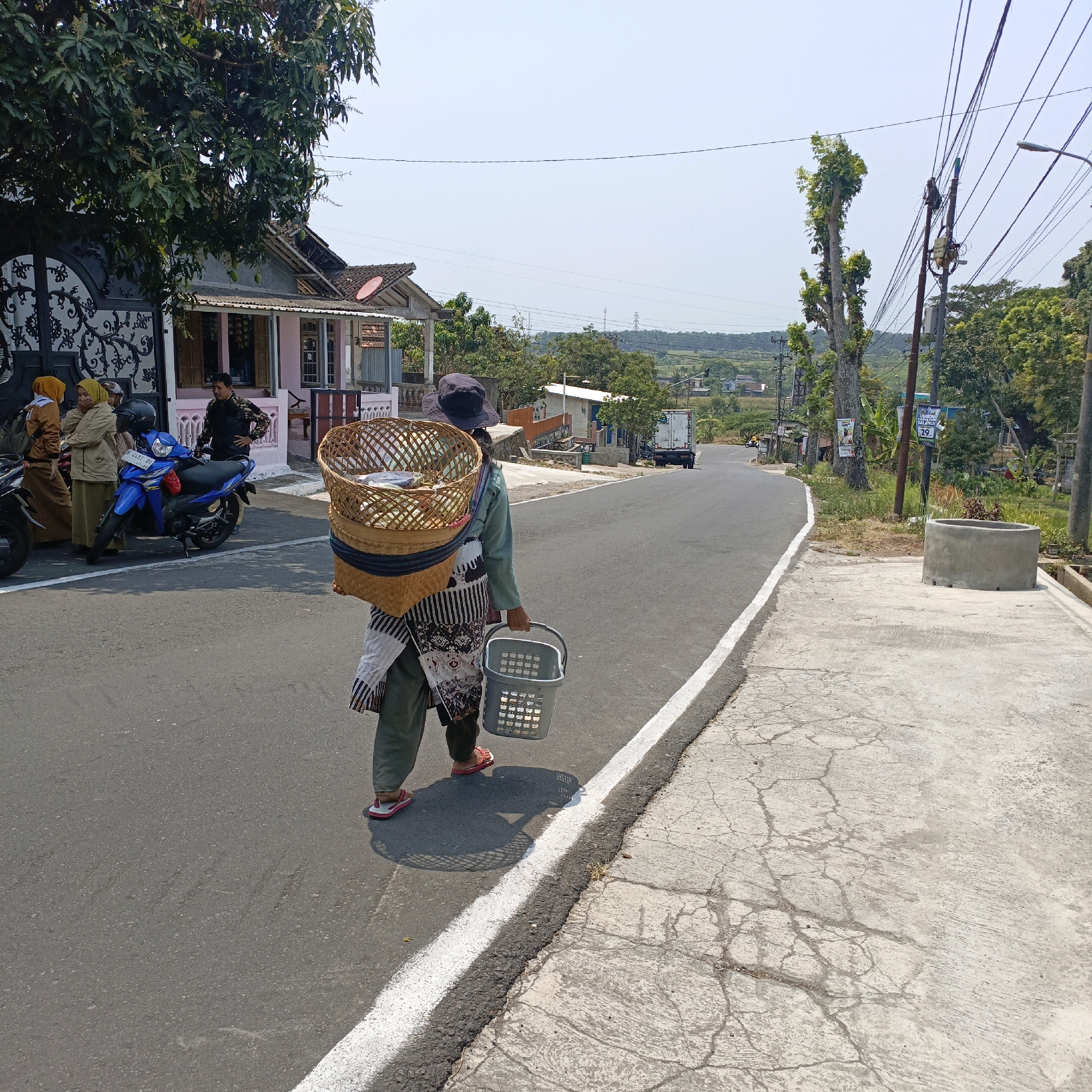 The image depicts a street scene in Salatiga, Indonesia, on a bright, sunny day. In the foreground, a person is walking away from the camera, carrying a large woven basket on their back and a gray plastic basket in their right hand. The person is wearing a green long-sleeved shirt, dark pants, a patterned apron or skirt, a hat, and red flip-flops. The woven basket on their back appears to contain various items, possibly goods for sale.
To the left of the street, a blue motorcycle is parked near a building with a pink facade and dark decorative fencing. Three individuals, two women in hijabs and a man, are standing near the motorcycle, appearing to be conversing.
The street itself is paved and has a white line marking the edge. To the right of the street, a cracked concrete sidewalk runs alongside. A cylindrical concrete structure, possibly a well or planter, is visible on the sidewalk.
In the background, the scene unfolds with more buildings, trees, utility poles with numerous wires, and a distant landscape of rolling hills under a clear blue sky. A white truck is visible driving on the road in the middle distance. The overall environment suggests a semi-rural or suburban area.