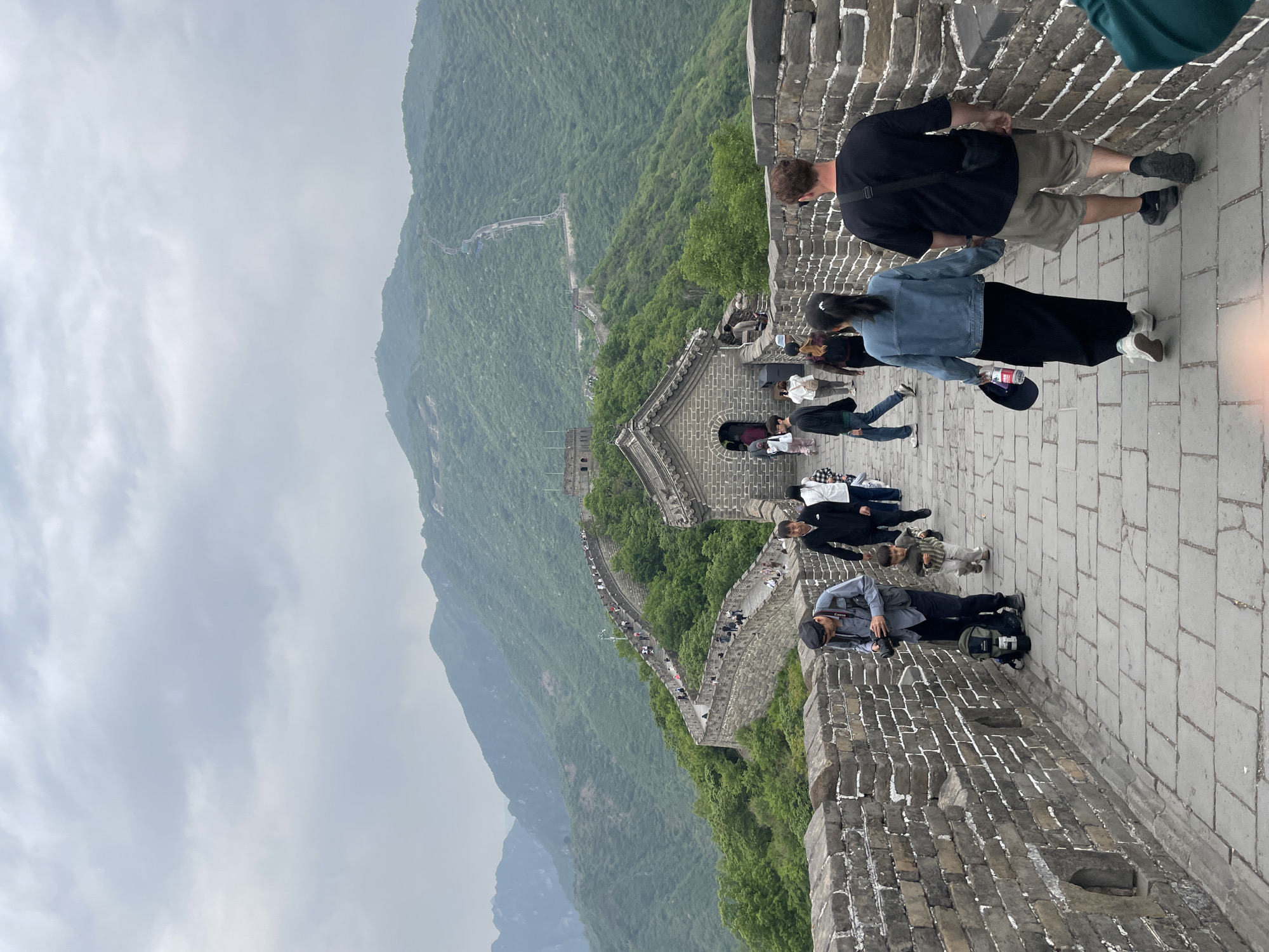 Captured at the Great Wall of China in the Huairou District of China, the image presents a high-angle perspective. A group of tourists is seen walking along the stone path of the wall. The wall itself extends into the distance, winding through a green, mountainous landscape, with a visible fortress-like structure along its path. The sky is overcast. The presence of tourists and the structure of the wall serve as focal points in the scene.