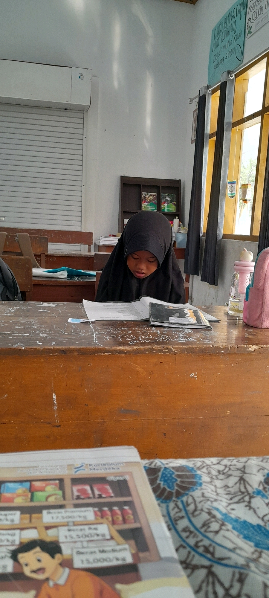 In a classroom in Rogojampi, Indonesia, a young girl wearing a black hijab is diligently studying at a wooden desk. Sunlight streams in from a window covered by yellow and black curtains, illuminating the room and casting shadows on the white walls. The girl's gaze is fixed on an open book on her desk, which also holds a black notebook and a pink water bottle. In the background, a shelf displays a few books, and rows of empty wooden desks are visible. A rolled-up white shutter covers one wall. In the foreground, another book with cartoon illustrations of products and prices is partially visible, resting on a patterned cloth. The overall scene depicts a typical school day, with a focus on the girl's engagement with her studies.