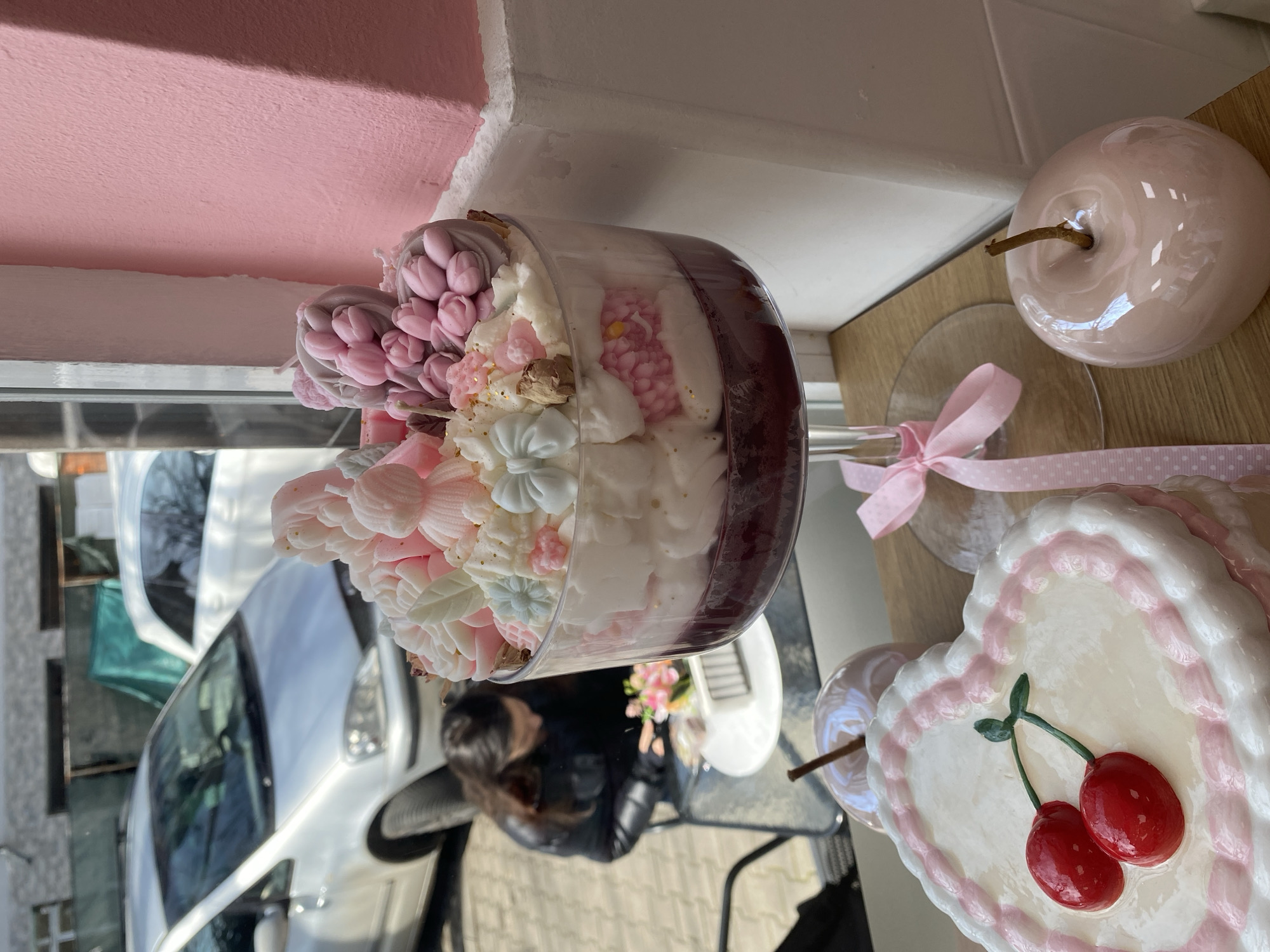 The scene depicts the interior of a shop, likely a patisserie or cafe, looking out onto a street in Stara Zagora, Bulgaria. In the immediate foreground, a large, clear glass trifle-style dessert or display item on a stemmed base dominates the center. This item features distinct layers: a dark reddish-purple base, followed by a thick layer of white cream, a pink textured layer containing small spherical elements, another white cream layer, and a top layer of white cream heavily adorned with decorative elements. These decorations include pale pink sculpted figures, white and light pink sculpted roses, light blue and pink bows, small golden sprinkles, and a cluster of small pink tulip-shaped forms on a grey base.
To the lower left, a heart-shaped white item, possibly a cake or decorative piece, is visible with pink piped icing around its edge and two prominent red cherry-like objects with green stems on its surface. To the lower right, a shiny, pale pink apple-shaped figurine with a brown stem rests on the surface. These items are situated on a light brown wooden surface, likely a window sill or counter. A pink ribbon with white polka dots is tied around the stem of the large glass display. A textured pink wall is visible to the right of the white window frame.
Through the window, an outdoor area is visible. A single individual with long, dark hair, wearing a black puffer jacket, is seated at a small, round, white table with a dark metal chair. The person is looking down, engaged with items on the table, which include a small floral arrangement with pink flowers and a white rectangular box. Two cars are parked outside: a silver or light grey minivan is partially visible on the left, and a white car is partially visible behind it on the right. The ground outside is paved, and further in the background, obscured structures with green coverings are visible under bright daylight.