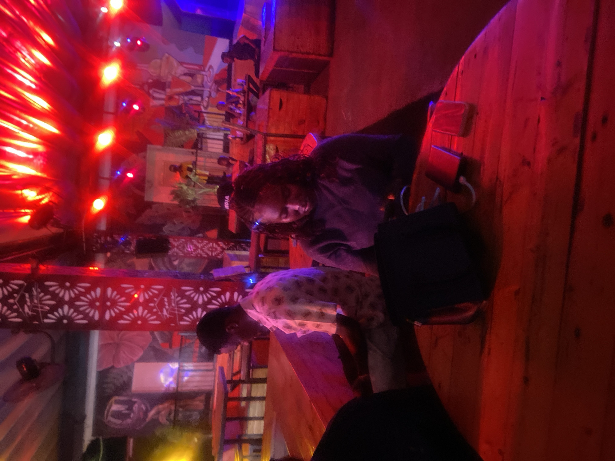The image depicts an indoor setting, likely a bar or lounge, bathed in the vibrant glow of red and blue stage lights. In the foreground, a wooden table is visible, with a dark bag and a power bank with its cord resting on it. To the left, a man in a patterned shirt is seated, looking down. To the right, a woman with braided hair, wearing a dark top, is also seated, looking downwards at the table. The background reveals more seating areas and what appears to be a DJ booth with a person visible. The walls are decorated with murals and patterned pillars. The overall atmosphere is dark, with the red lighting dominating the scene and creating a lively, possibly nocturnal, ambiance. This scene is set in Howayi, Nigeria.