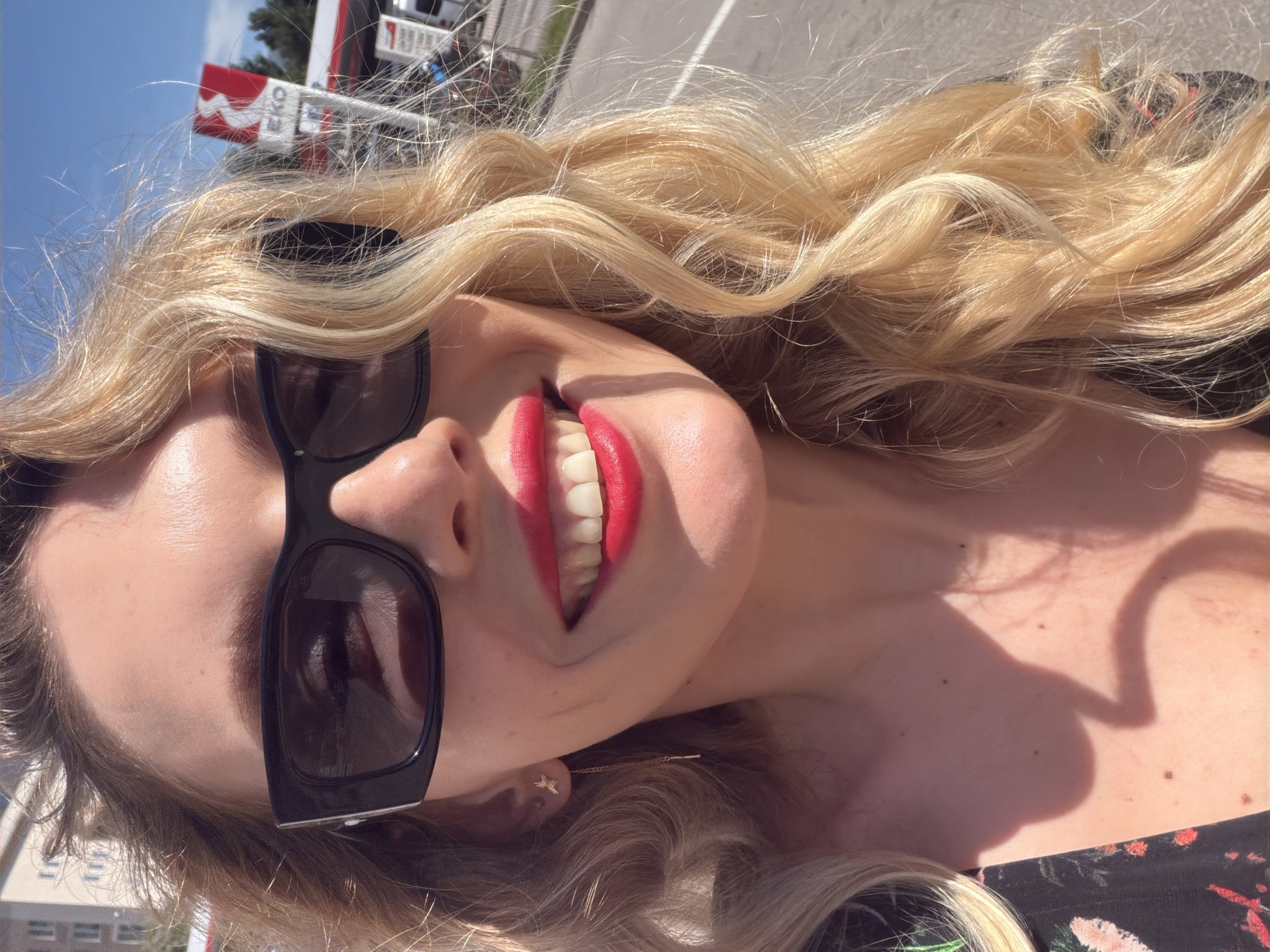 A close-up, tilted portrait captures a person, likely a woman, outdoors on a bright, sunny day in what appears to be Budva, Montenegro. The subject is smiling broadly, revealing bright white teeth and vibrant red lipstick. She is wearing dark, rectangular sunglasses that cover her eyes, with a reflection of the sky and surroundings visible in the lenses.
Her long, wavy, blonde hair cascades around her face and shoulders, catching the sunlight and showing individual strands. A small, delicate, star-shaped earring is visible on her right ear. Strong sunlight casts distinct shadows across her neck and upper chest, indicating a high sun position. Part of her attire, featuring a dark fabric with a floral pattern in red and green hues, is visible in the bottom right corner.
In the background, under a clear blue sky, elements of what looks like a gas station or commercial area are visible. A white sign with red accents can be seen, prominently displaying the text "EKO". The overall mood conveyed is one of joy and brightness, likely a vacation or leisure moment.