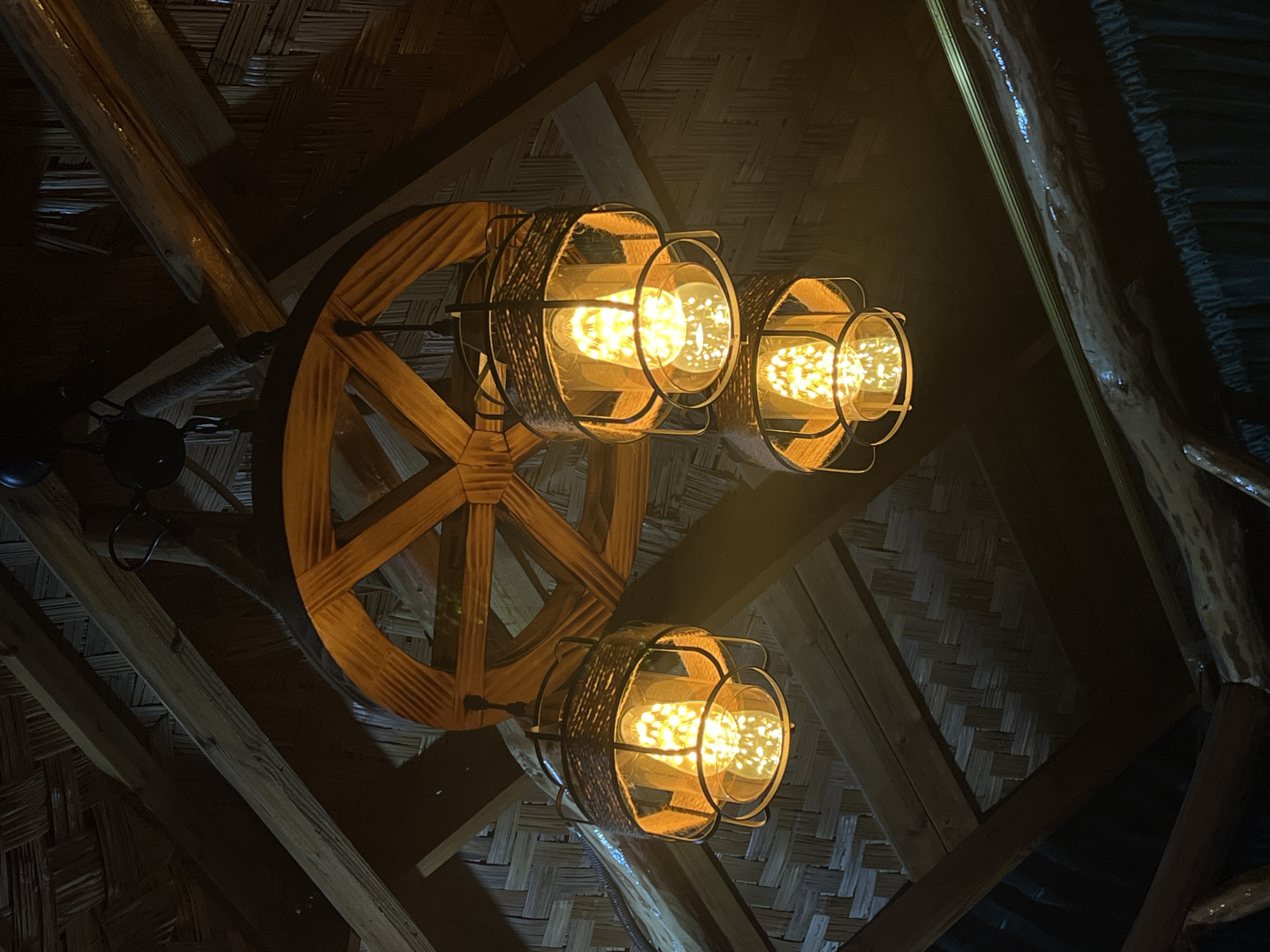 This image shows a rustic chandelier with three Edison-style bulbs hanging from a wooden ceiling. The chandelier is suspended from a wooden wheel-like structure, and the lampshades are made of woven material and metal. The ceiling is constructed from wooden beams and woven mats, giving it a natural, earthy feel. The lighting is warm and ambient, creating a cozy atmosphere. The scene appears to be indoors, likely in a restaurant or cafe with a traditional or rustic design. The location is specified as being in the city of Namangan City, Uzbekistan. There are no people visible in the image.