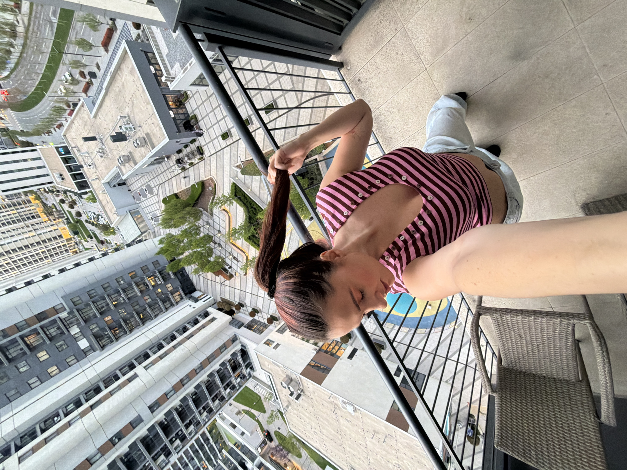 A woman with her hair in a ponytail is standing on a balcony overlooking a cityscape. She is wearing a pink and brown striped top and light-colored pants. Below the balcony are buildings with a mix of apartments and offices. There is a plaza with landscaping including trees and grass. The image was taken in Belgrade, Serbia.