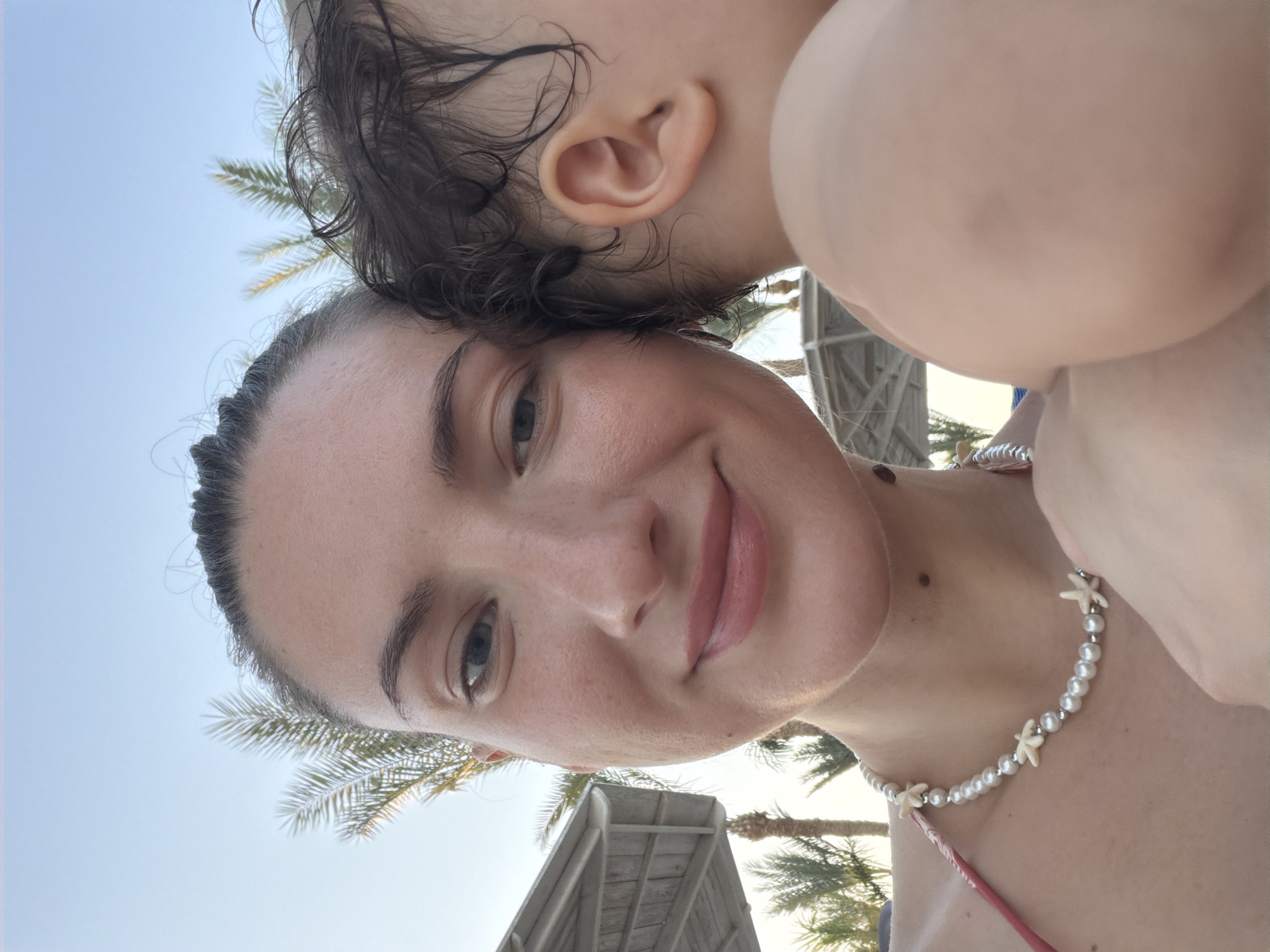A selfie is taken outdoors, with a clear sky and palm trees in the background, suggesting a location like Sharm El Sheikh, Egypt. The image features a woman in the foreground, holding a baby close to her. The woman is looking towards the camera and smiling. She is wearing a bikini top and a pearl necklace with starfish charms. The baby's ear is visible, along with part of its shoulder and head. In the background, parts of beach umbrellas and palm trees are visible. The lighting suggests it is daytime, possibly late afternoon.