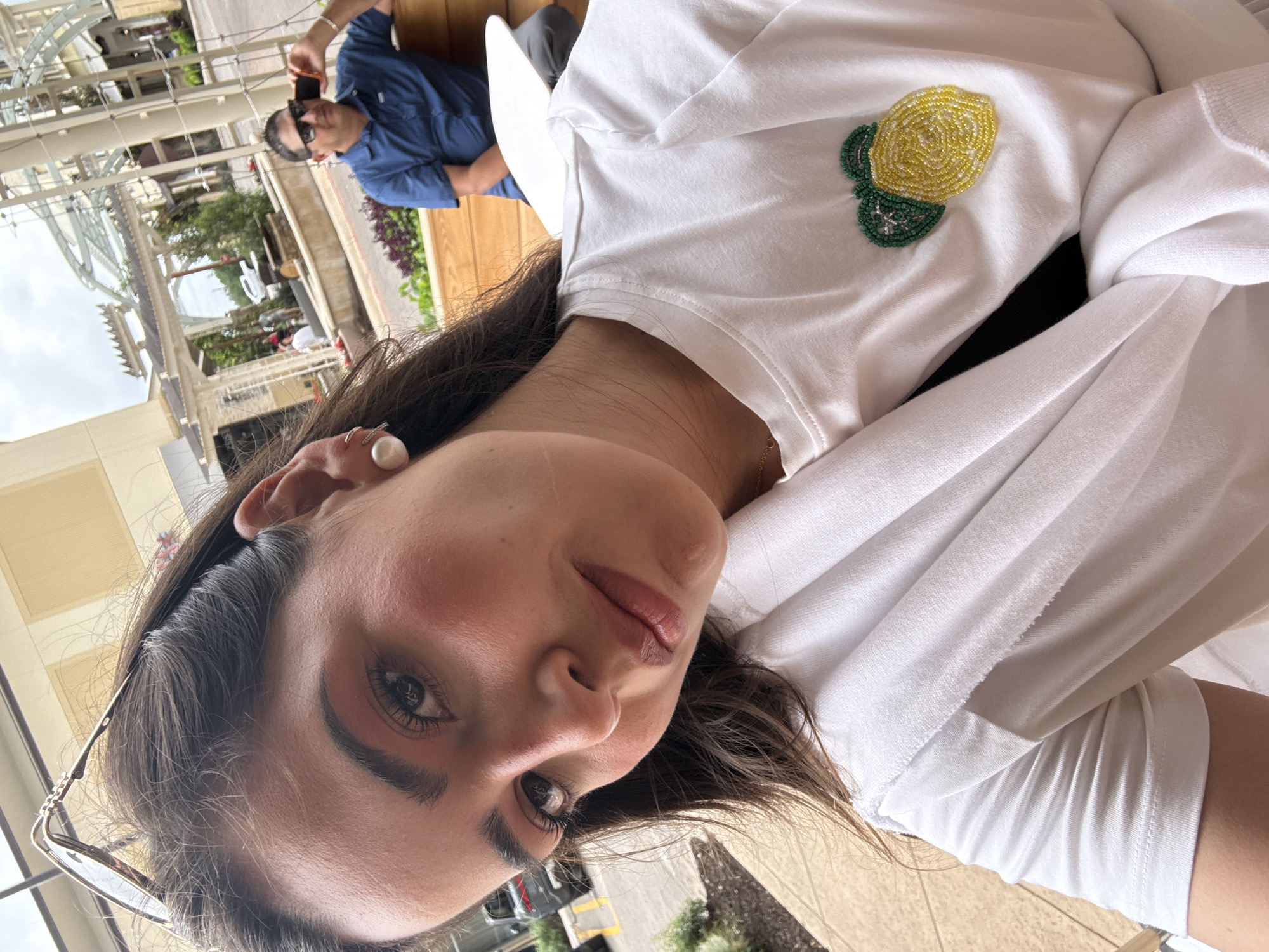 The image shows a woman in a white t-shirt with a beaded lemon pin on the front. She is wearing pearl earrings and glasses are tucked into her hair. In the background, a man wearing a blue shirt and sunglasses is holding a phone. The scene appears to be an outdoor seating area, with a metal pergola structure and greenery visible. The location is likely in San Antonio, United States of America.