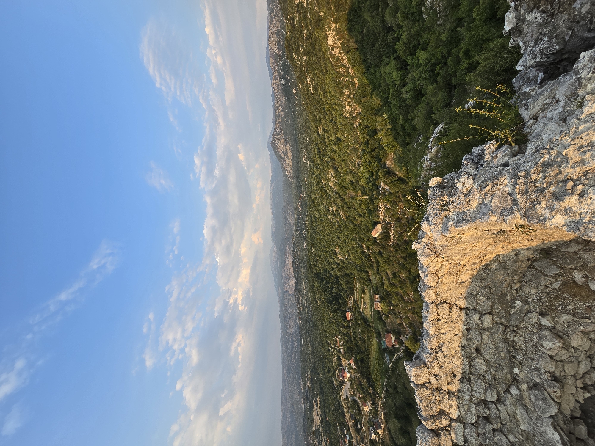 The image depicts a scenic view from the ruins of a fortress in Medun, Montenegro. In the foreground, weathered stone walls of a circular structure, likely part of the fortress, are visible. These stones are rough and appear to be aged, with patches of sunlight illuminating their texture. Beyond the ruins, a lush green valley stretches out, dotted with small houses and vineyards. The landscape is characterized by rolling hills covered in dense vegetation, transitioning into more rugged, mountainous terrain in the distance. The sky above is a clear blue, with scattered wispy clouds, suggesting it is either early morning or late afternoon, as indicated by the warm, directional sunlight casting shadows across the scene. The overall impression is one of historical significance combined with natural beauty.
