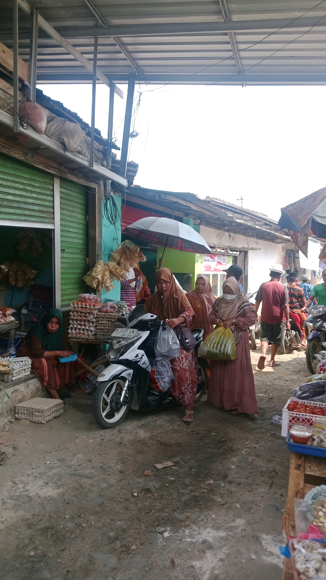 This image depicts a street market scene in Waru, Indonesia. The foreground shows a dirt path lined with market stalls. In the center, a white scooter is parked, with several women in modest clothing around it. One woman, wearing a brown hijab and dress, is interacting with the scooter. To the left, another woman in a green hijab and striped brown dress sits near stacks of egg cartons. Behind them, a stall has a green rolling shutter. Several people are walking in the background, some on motorcycles. The market appears to be covered by a metal roof structure. The atmosphere is that of a busy, local marketplace.