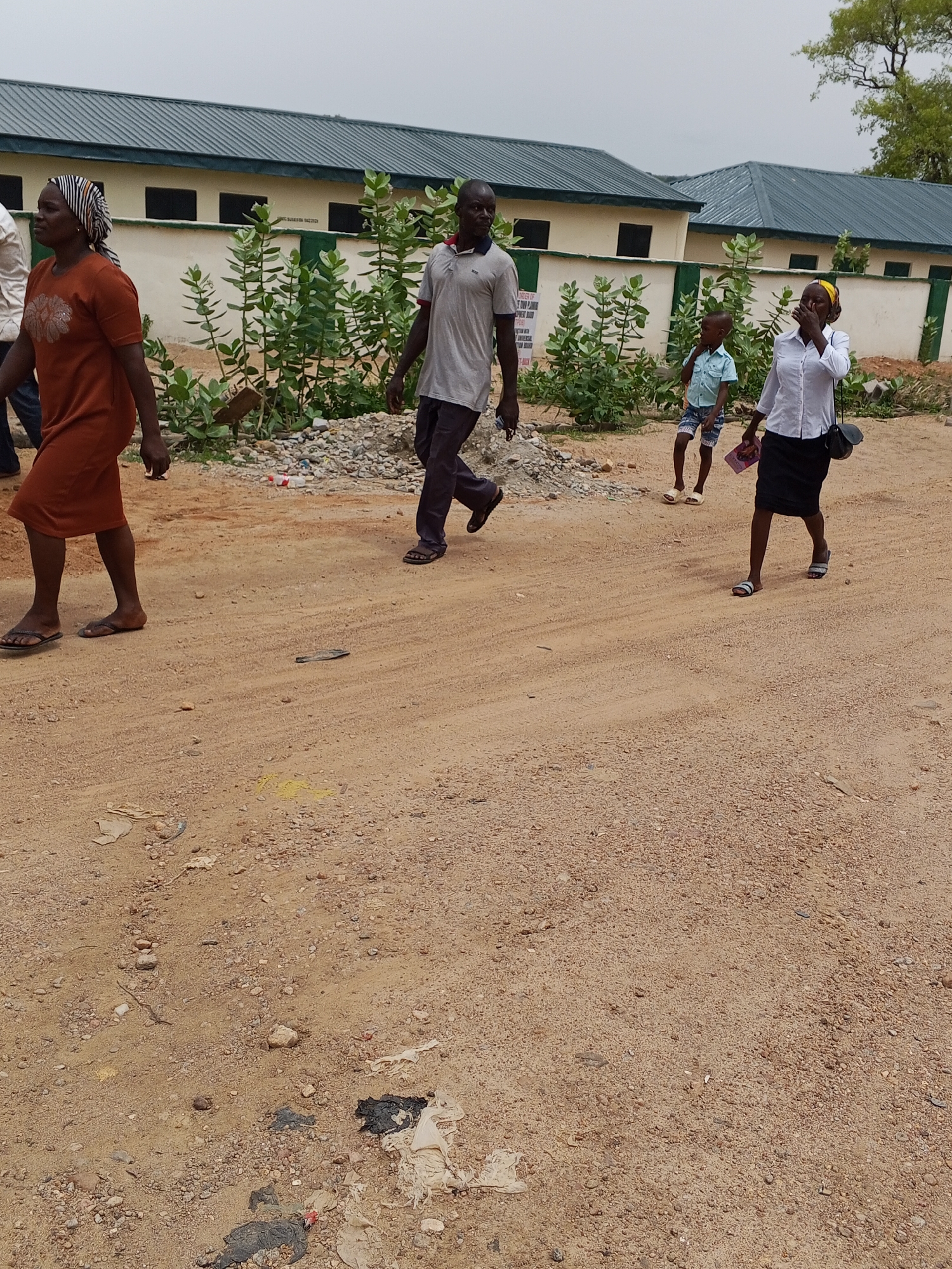 A medium-shot image depicts four people walking on a dirt road in Otokiti, Nigeria, during daylight. In the foreground, a woman in a brown dress and head covering walks to the left. Next to her, a man in a grey shirt and dark pants walks in the same direction. Further away, a young boy in a light blue shirt and shorts walks alongside a woman in a white blouse and black skirt, both moving to the right. Behind them, a low, light-colored building with a green roof is visible. The area adjacent to the building contains greenery. The ground is dirt, littered with debris. The sky is overcast.