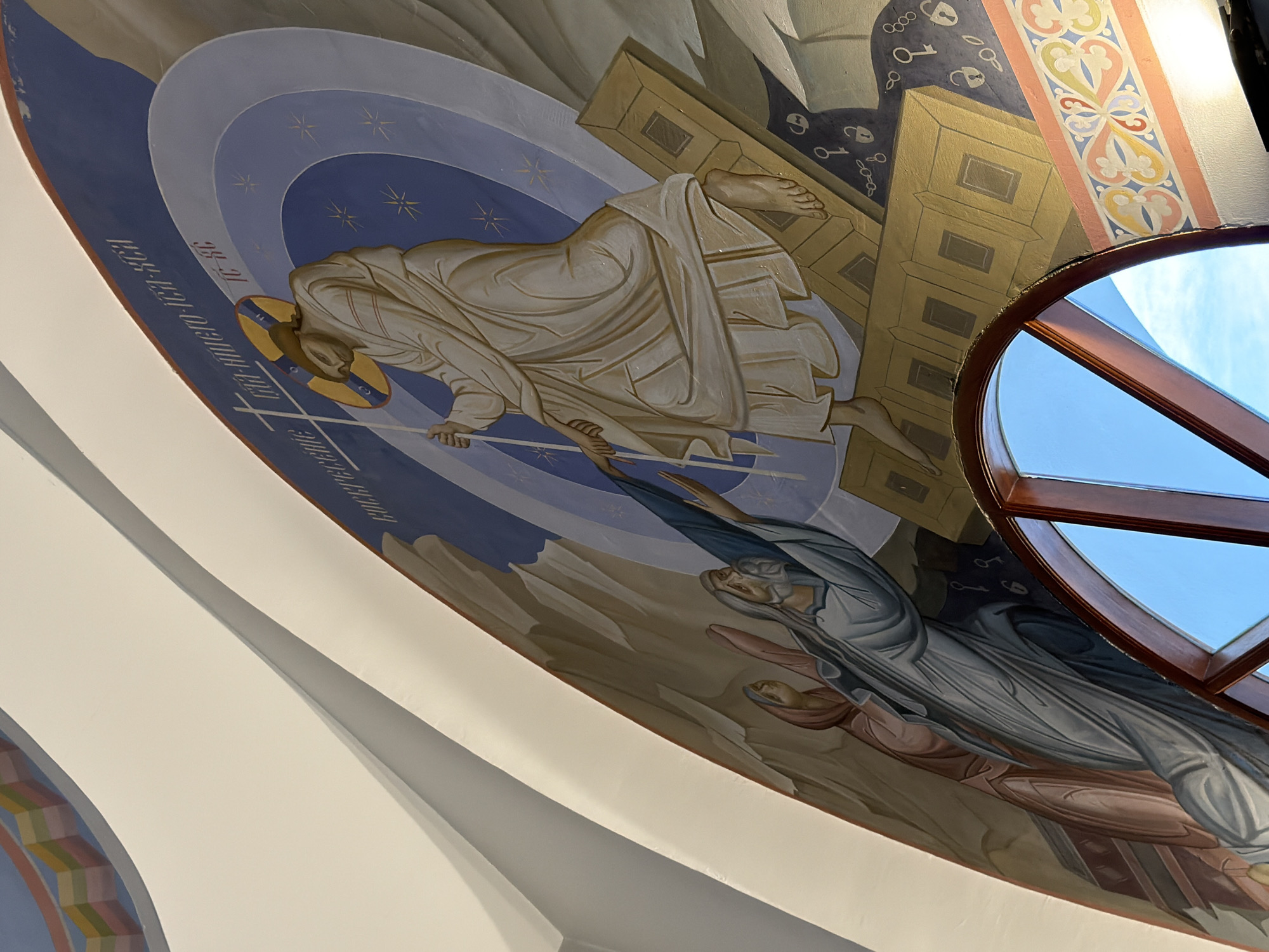 This is a close-up, angled shot of a fresco depicting a religious scene. The fresco is painted on a curved surface, likely the ceiling or an arched wall of a building. In the center, a figure, presumably Jesus Christ, is depicted rising with a halo and a cross. He is stepping onto a golden structure, possibly representing a tomb or ascension platform. Below and to the left, two figures in flowing robes are reaching up towards the central figure. One figure, appearing older with a beard, is in a blue robe, and the other, a woman in a pinkish robe, is partially visible. The background transitions from rocky, mountainous terrain to a celestial scene with blue hues and star-like elements. There are also visual elements of chains and locks depicted in a darker blue area, suggesting liberation. The fresco is rendered in a traditional Byzantine or Orthodox iconographic style. The bottom of the image shows a segment of a wooden-framed circular window with a bright blue sky visible through it, suggesting the fresco is located in an area with natural light. Decorative borders with floral motifs are visible along the bottom edge of the fresco. The overall impression is one of resurrection and divine triumph. The location of this fresco is within a religious building in Ko Samui District, Thailand.