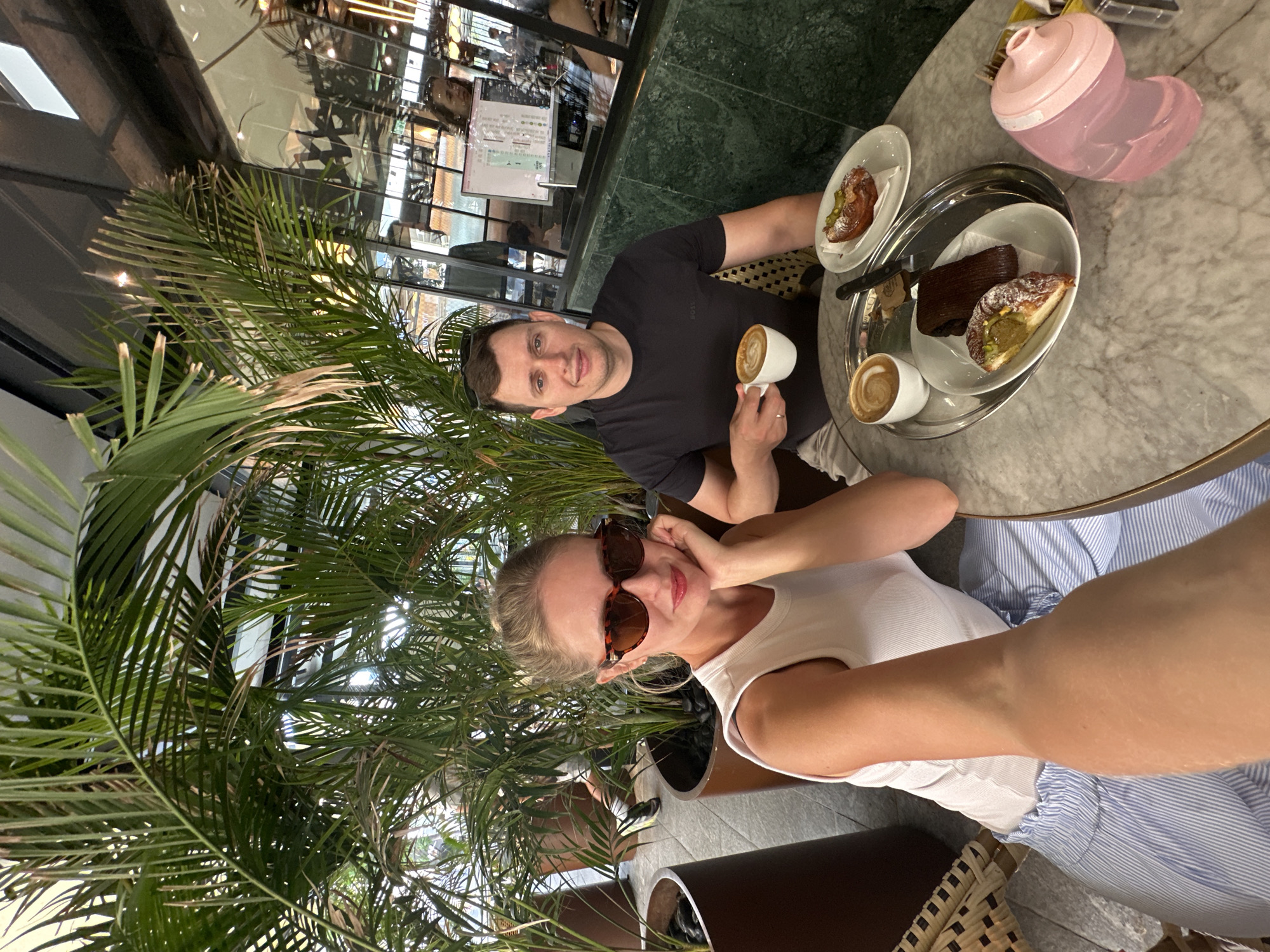In Herzliya, Israel, a man and a woman are seated at a marble-topped table in a cafe, surrounded by lush green palm plants. The woman, wearing sunglasses and a white top, is in the foreground, resting her chin on her hand. The man, in a dark t-shirt, holds a cup of coffee. On the table are two more cups of coffee, a tray with pastries and chocolate, and a pink baby bottle. The cafe's interior, visible through reflections in the glass windows behind them, suggests a modern and airy setting with patrons in the background.