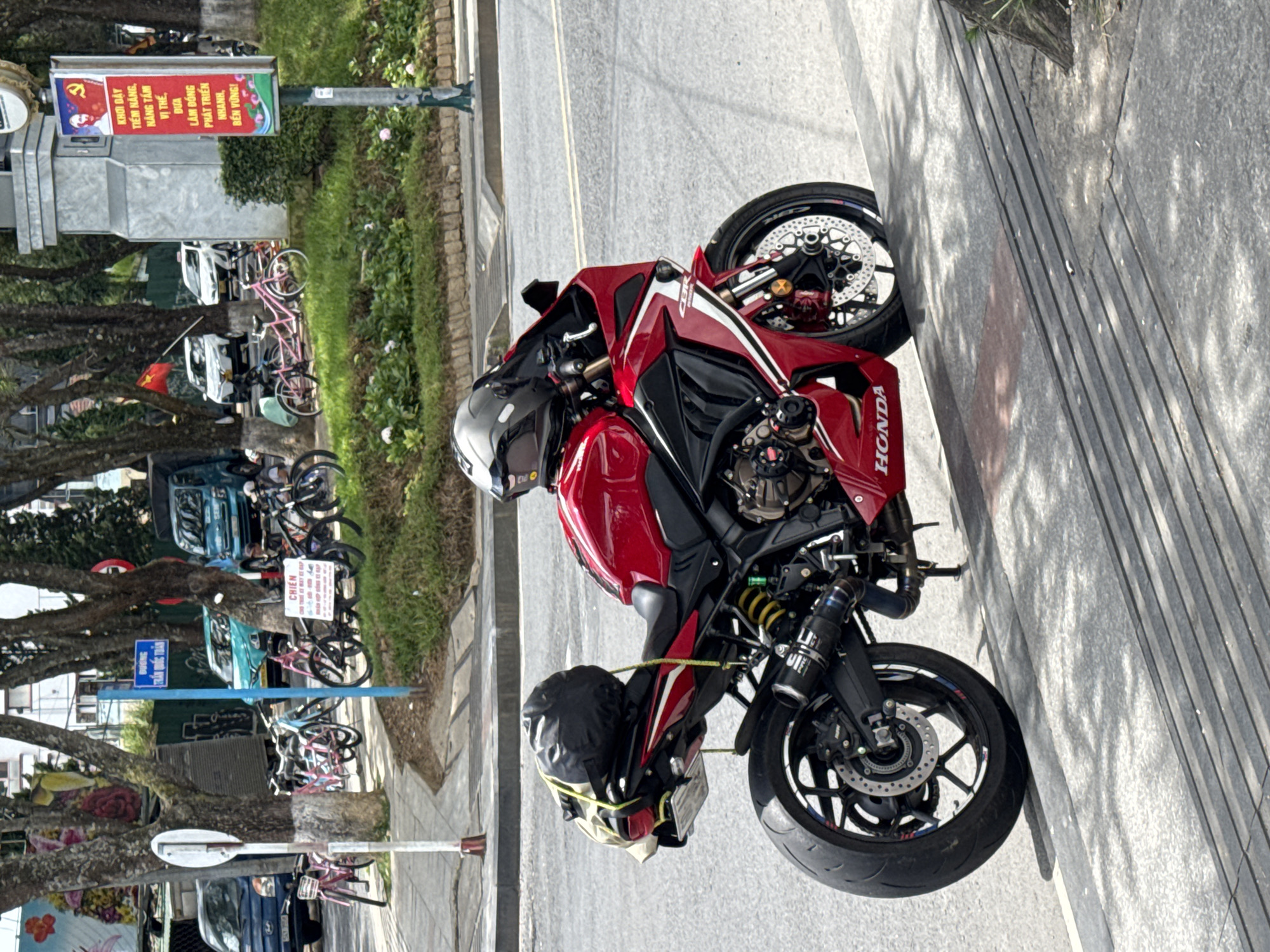 This media file depicts a red Honda CBR motorcycle parked on a sidewalk next to a street. The motorcycle is the primary subject, and it appears to be a sunny day, casting shadows from trees onto the scene. To the left of the motorcycle, a row of trees lines the street, with some parked bicycles and a blue truck visible in the background.
A prominent sign with Vietnamese text is visible, featuring a communist-style imagery and slogans that translate to "Arouse potential, elevate position, build Lam Dong's rapid and sustainable development!". This suggests the location is likely in Vietnam, specifically in the Lam Dong province, and the sign indicates a public or political context. Another smaller blue sign with Vietnamese text is also present. The overall setting appears to be an urban or semi-urban environment, possibly a street in Da Lat, Vietnam, given the context clues. There are no people clearly visible in the frame, but the presence of the motorcycle and bicycles suggests this is a public area. The motorcycle is equipped with a black bag strapped to the back, possibly for carrying items. The image is taken from a slightly elevated angle, looking down towards the motorcycle and the street.