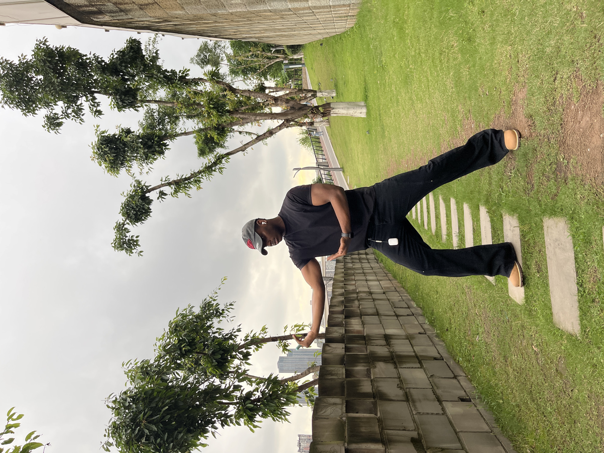 The photograph depicts a man in Yuzhong District, China, standing in a park. He is wearing a black t-shirt, black pants, tan shoes, and a baseball cap. He is positioned on a low wall made of gray stone blocks, reaching towards a tree with green leaves. The background features a grassy area with stepping stones, trees, and a cloudy sky. A modern skyline with skyscrapers is visible in the distance.