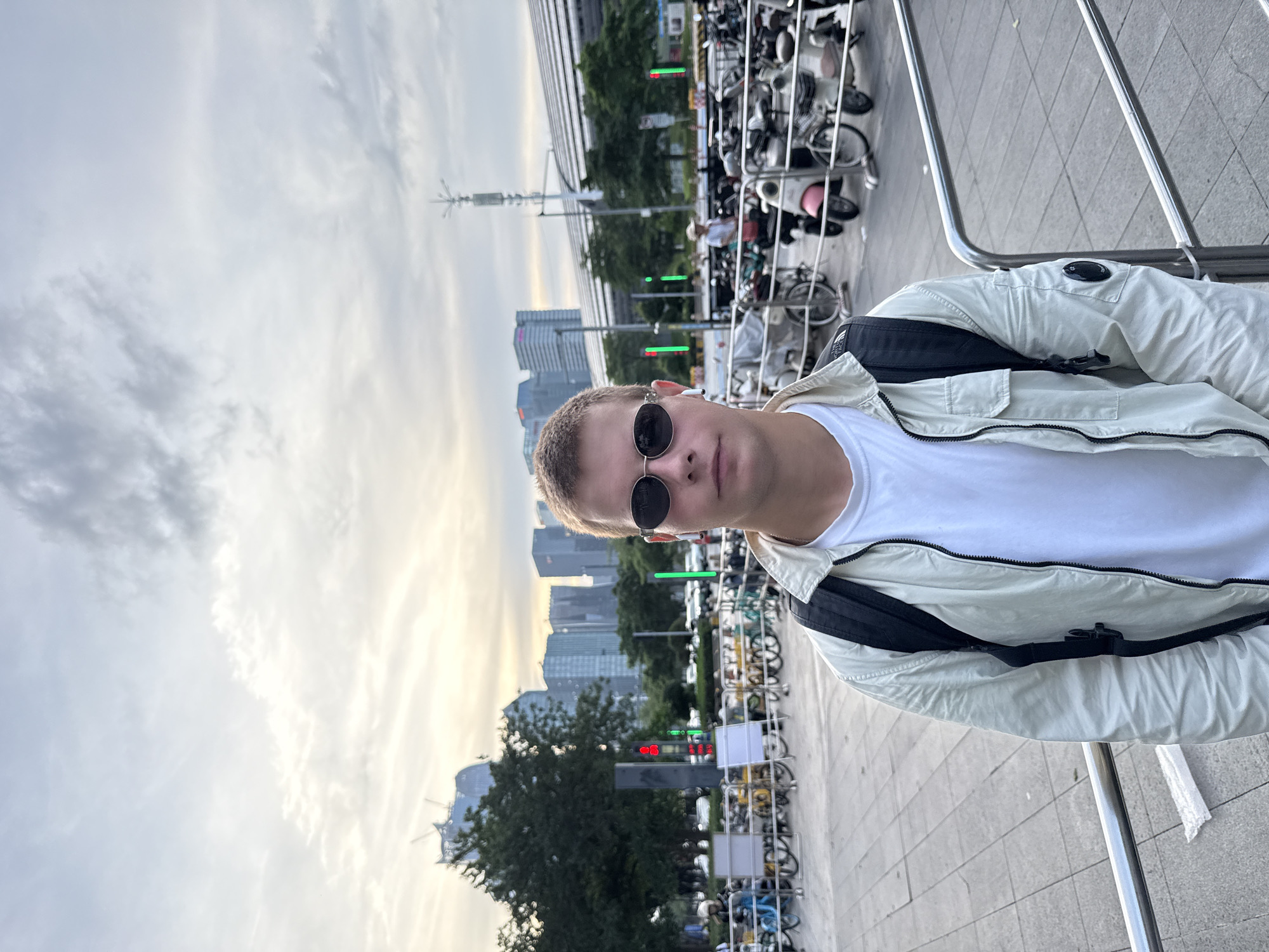 A young man stands in Haizhu District, China, wearing a white t-shirt, light-colored jacket, and black sunglasses. He is wearing a black backpack. Behind him are parked bicycles, a row of trees, and buildings silhouetted against the sky. A metal railing is to his right. The sky is cloudy.
