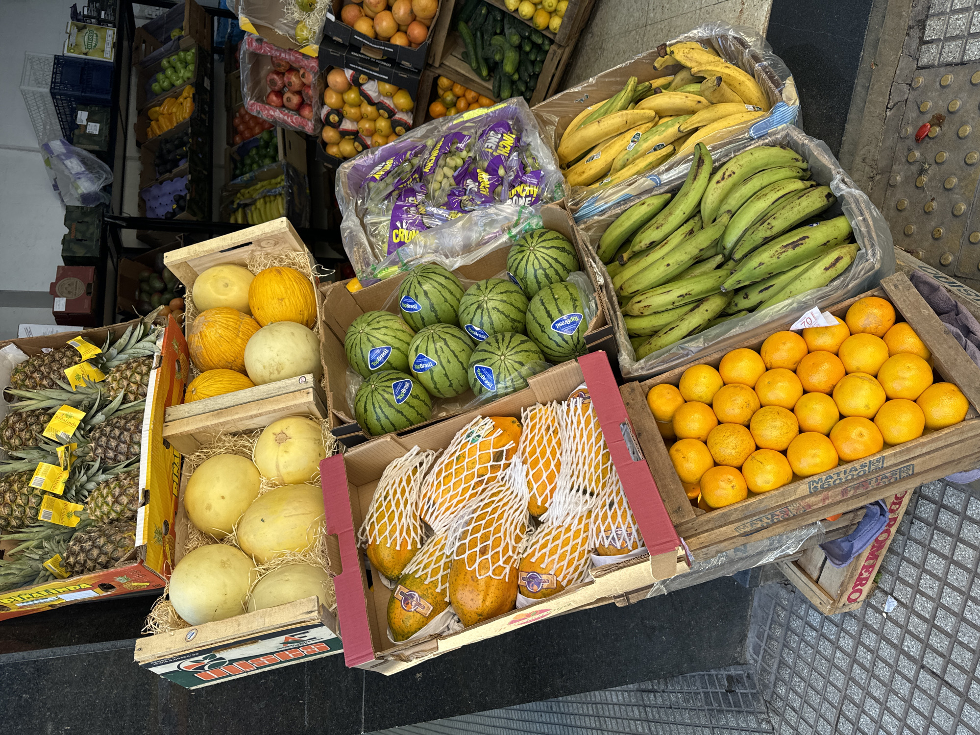 An eye-level, medium-shot depicts various crates of fruit displayed on a sidewalk in Buenos Aires, Argentina. The fruits, arranged in cardboard and wooden crates, include pineapples, honeydew melons, yellow melons, mini watermelons, papayas wrapped in protective netting, bunches of purple grapes, yellow bananas, green plantains, and oranges.
The pineapples and melons are in wooden crates, while other fruits are in cardboard boxes with plastic lining. The mini watermelons and papayas are branded with labels "Top Brasil" and the grape bunches are “CRUNCH TIME GRAPES”. The crates are arranged seemingly randomly along a black wall and the sidewalk, which is patterned with a cross-hatch grid design. The fruits are fresh.
Some boxes show text like “Piñalinda” and “MATIAS S.A.”
The location appears to be a street vendor's display outside a store. The sidewalk is on the right and the black wall on the left.