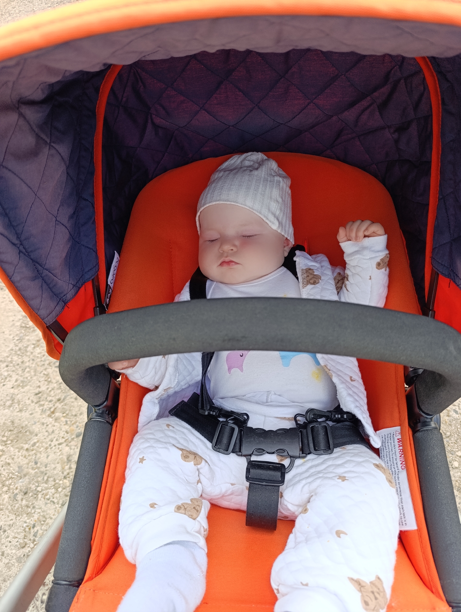 A baby is captured sleeping peacefully in a vibrant orange stroller. The infant wears a white ribbed beanie and a white quilted outfit patterned with small brown teddy bears and stars, layered over a white long-sleeved top featuring a pastel unicorn and rainbow graphic. The baby's eyes are closed, and their small hands are gently fisted, resting near their chest. They are secured by a black safety harness with a large buckle.
The stroller has a bright orange padded seat and lining, contrasting with the dark grey or black quilted canopy lining, which is edged with an orange trim. A black padded bumper bar extends across the front of the baby. The setting appears to be outdoors during bright daylight, with a light-colored, possibly gravel or paved, ground surface visible beneath and around the stroller. The overall mood is serene and calm, suggesting a comfortable rest for the baby during an outing.
Visible text on a white warning label attached to the harness strap reads: "AWARNING. Audible click must be heard in order to determine that each lock is in proper position. To ensure proper locking sngae, chack to see that diserne seat/bassinet/Bugabe disengaged tabe use." A small white tag with "bugaboo" (partially visible as "bigoba") is also present inside the canopy. The photo was taken in Chaikovsky, Russia.