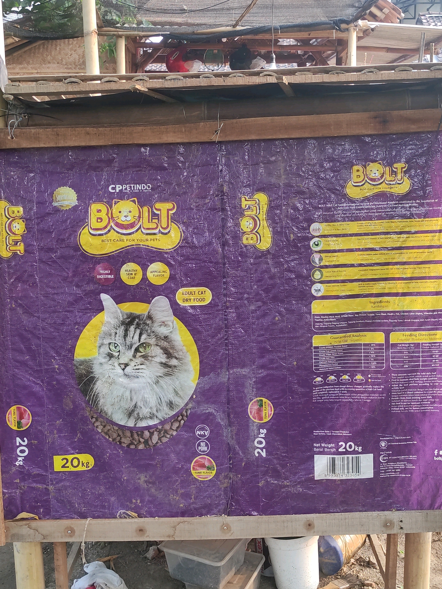 The image shows a large, purple plastic bag of "Bolt" brand cat food, likely displayed outdoors at a stall or market in Tiudan, Indonesia. The bag features a prominent image of a cat's face, the brand logo, and nutritional information. The bag is attached to a wooden structure, possibly a temporary display stand. The background reveals a glimpse of what appears to be a rural or semi-rural environment, with bamboo poles, roofing materials, and some vegetation. There are no people clearly visible in the image. The overall scene suggests a commercial setting where pet food is being sold.