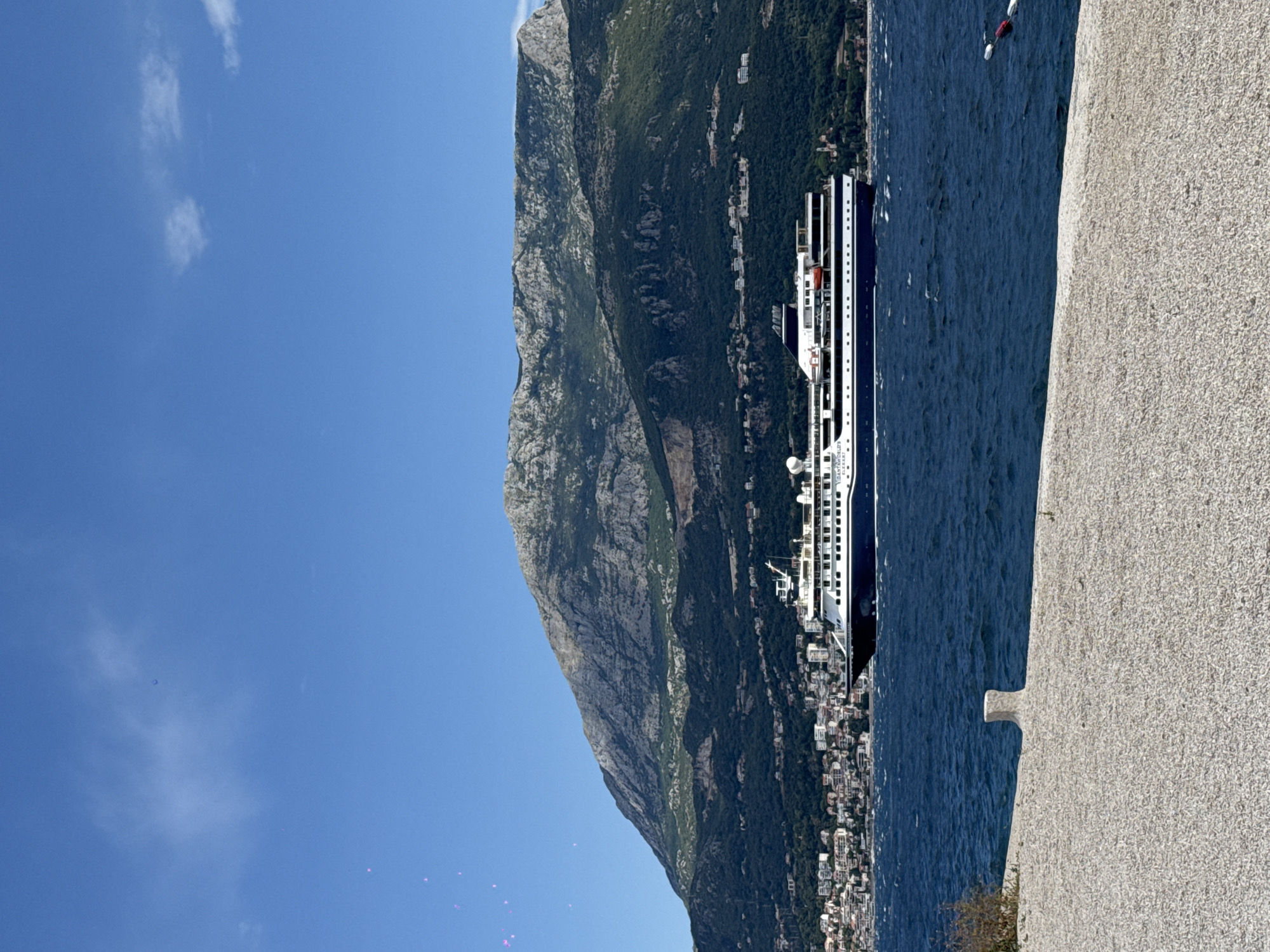 An eye-level, medium outdoor shot captures a large white and dark blue cruise ship sailing on a choppy, deep blue sea. The ship is positioned in the middle-right of the frame, moving from left to right. Behind the ship, a densely populated coastal town with white and beige buildings is visible, nestled at the foot of a large, imposing mountain covered in lush green trees and rocky grey patches. The sky above is a clear, bright blue with a few wispy white clouds. In the foreground, a textured, pale grey surface, possibly a quay or walkway, occupies the bottom third of the image. A single, small, off-white bollard stands on this surface. The overall scene suggests a sunny day, likely in a Mediterranean coastal region.