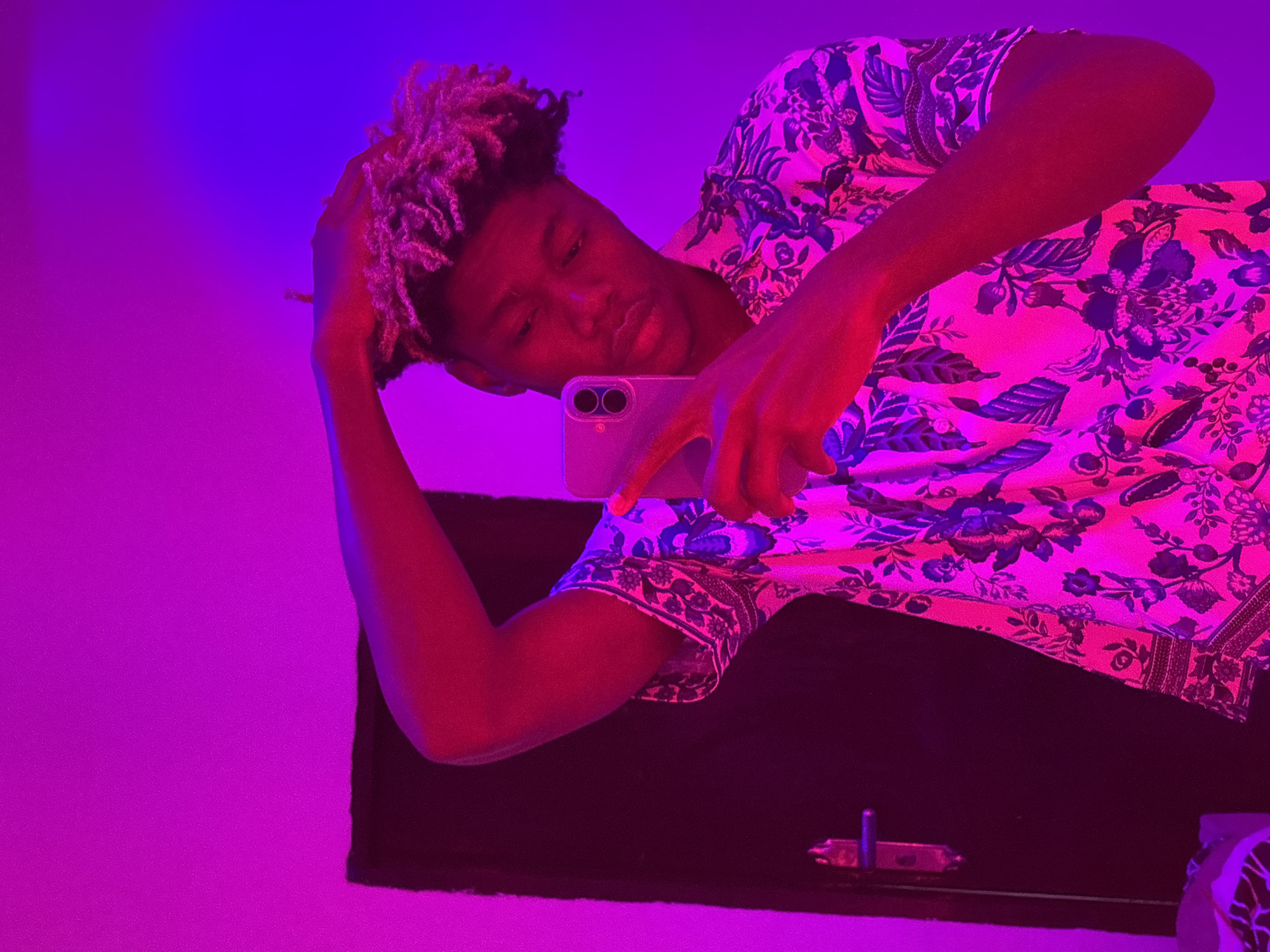 A young, Black male is seen in a self-portrait style photograph, taken in a room with purple and pink lighting. He has light brown, short, textured hair. He is wearing a white shirt with a blue floral print. He is holding a smartphone in his right hand, ostensibly taking the picture. The background is a solid purple color on the left and a dark surface, possibly a table or desk, is visible below him. Part of a white wall or surface is visible in the bottom right corner. There is no evidence in this image to identify the location as Igboefon, Nigeria.