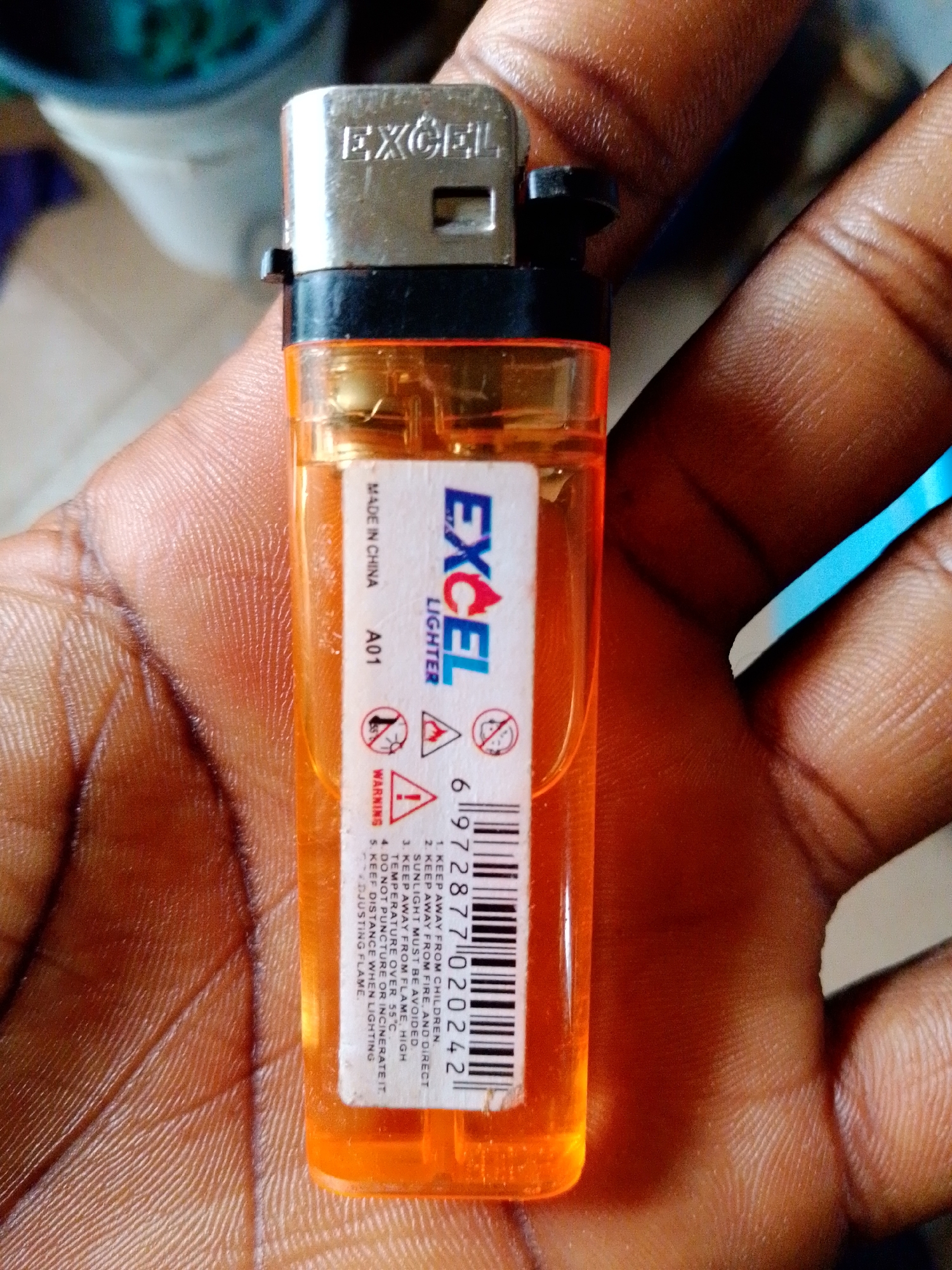 A close-up, eye-level shot shows a person's hands holding an orange Excel brand disposable lighter. The lighter has a silver top with the word "EXCEL" debossed on it, and a black plastic component that houses the igniter mechanism. The transparent orange body of the lighter reveals the internal workings and the fuel level. A white label is affixed to the body of the lighter, displaying the "EXCEL LIGHTER" logo, the model number "A01", a barcode, and a list of warnings in black text. Some of the warnings include "KEEP AWAY FROM CHILDREN," "KEEP AWAY FROM FIRE AND DIRECT SUNLIGHT MUST BE AVOIDED," and "TEMPERATURE OVER 55 C." The hands holding the lighter are dark-skinned, with visible fingerprints and texture on the skin. The background is blurred, but appears to be an outdoor or indoor setting with muted colors. The lighting is natural, casting soft shadows. No specific location within a city is discernible.