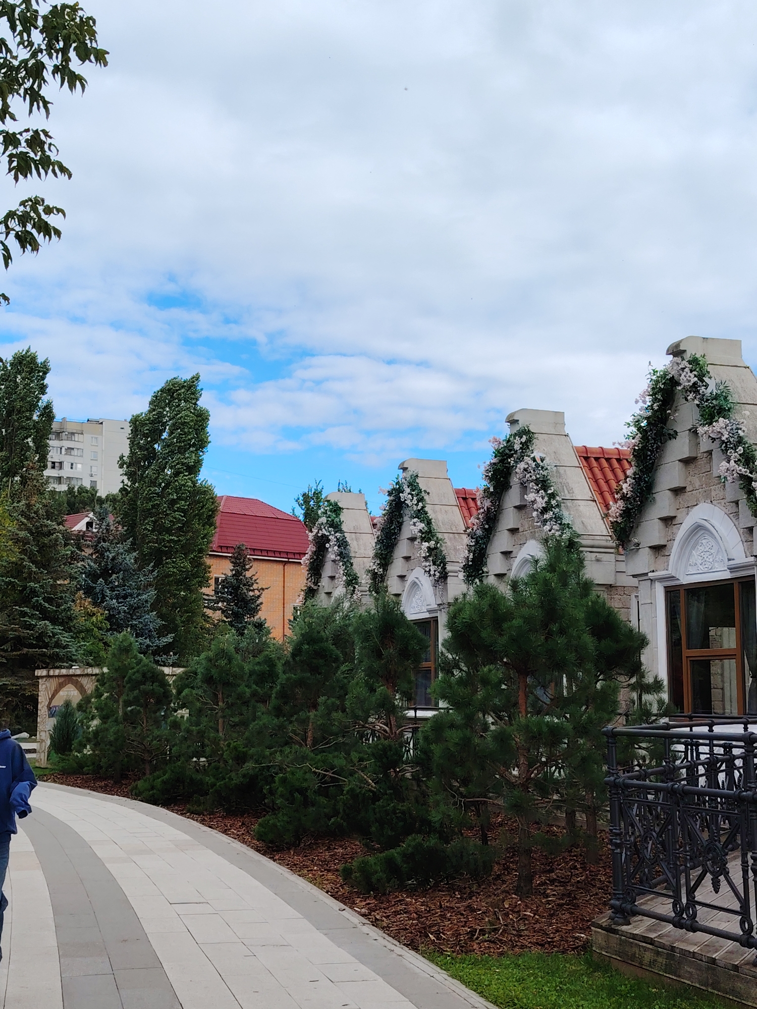 The image shows a row of architecturally unique buildings with pointed roofs, decorated with artificial floral garlands. The buildings appear to be a single structure designed to resemble multiple dwellings, possibly a hotel or a themed attraction. In the foreground, a paved walkway curves to the left, with a person in a dark blue hooded jacket and jeans walking away from the camera. To the right of the walkway, there is an ornate black metal railing and some greenery. Behind the buildings, trees and a taller apartment building are visible, indicating an urban or suburban setting in Zelenograd, Russia. The sky is partly cloudy.