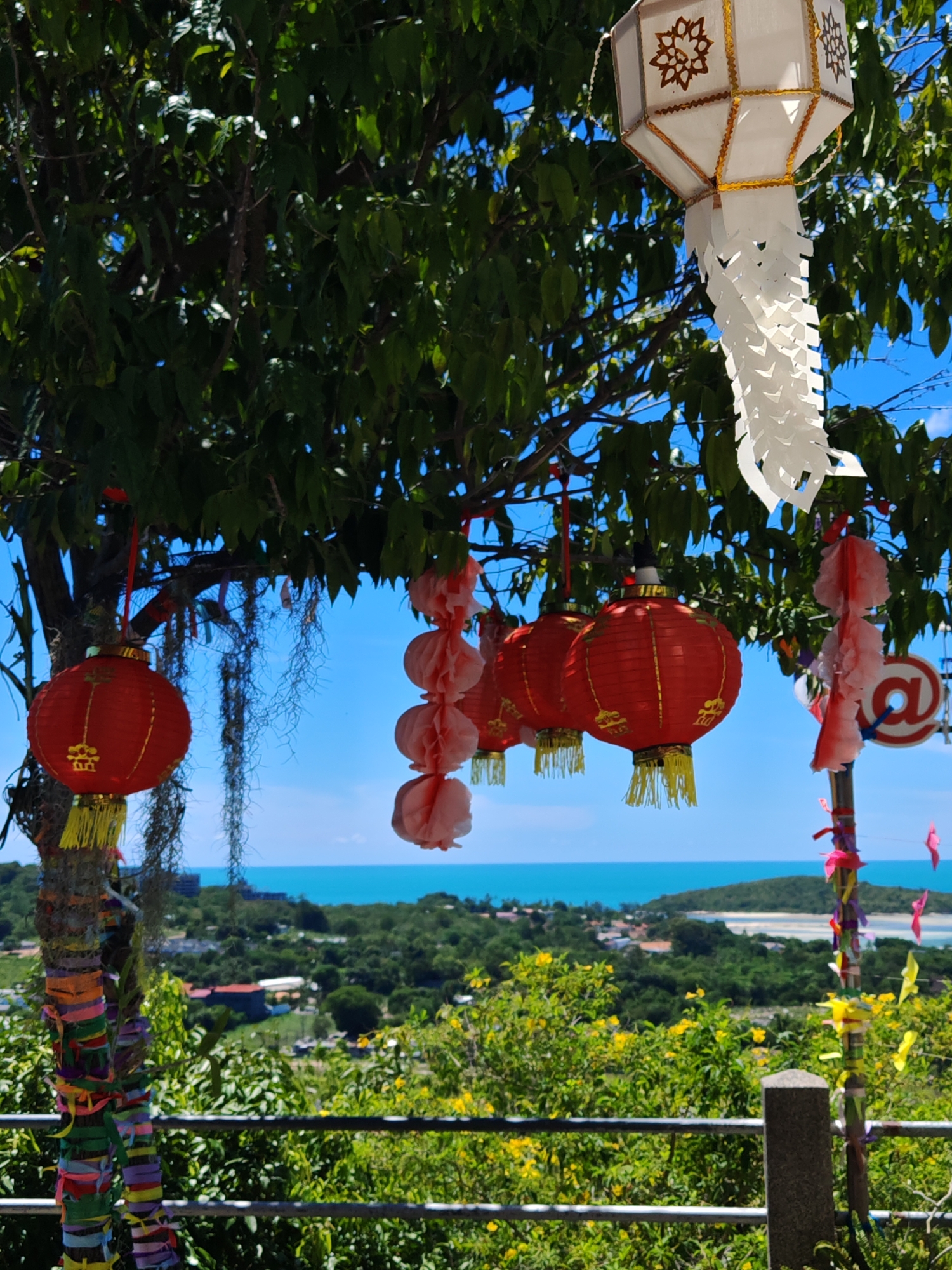 The image captures a scenic view of Baan Bang Rak, Thailand, with a focus on decorative lanterns hanging from a tree in the foreground. A large white lantern with ornate cutouts and gold trim is suspended from a branch, along with several smaller red lanterns adorned with gold embellishments and yellow tassels. Pink paper decorations are also visible, as well as thin, dried strands of vegetation hanging from the tree. The tree itself is lush and green, with a textured trunk wrapped in colorful ribbons.
Beyond the tree, a vibrant turquoise ocean stretches out under a clear blue sky. The coastline is dotted with buildings and lush green foliage, suggesting a coastal town or resort area. The overall atmosphere is bright and sunny, with the natural beauty of the landscape complemented by the festive decorations.