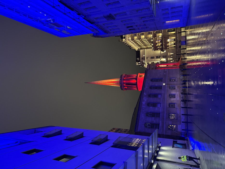 An urban nightscape is captured from a low angle, presenting buildings illuminated in vibrant colors. A prominent spire, bathed in red light, pierces the dark sky. Flanking this are buildings with blue and yellow lighting schemes, suggesting a modern, possibly artistic, urban environment. The ground appears wet, reflecting the intense colors, enhancing the dramatic effect. A figure in a yellow vest is visible at the bottom.