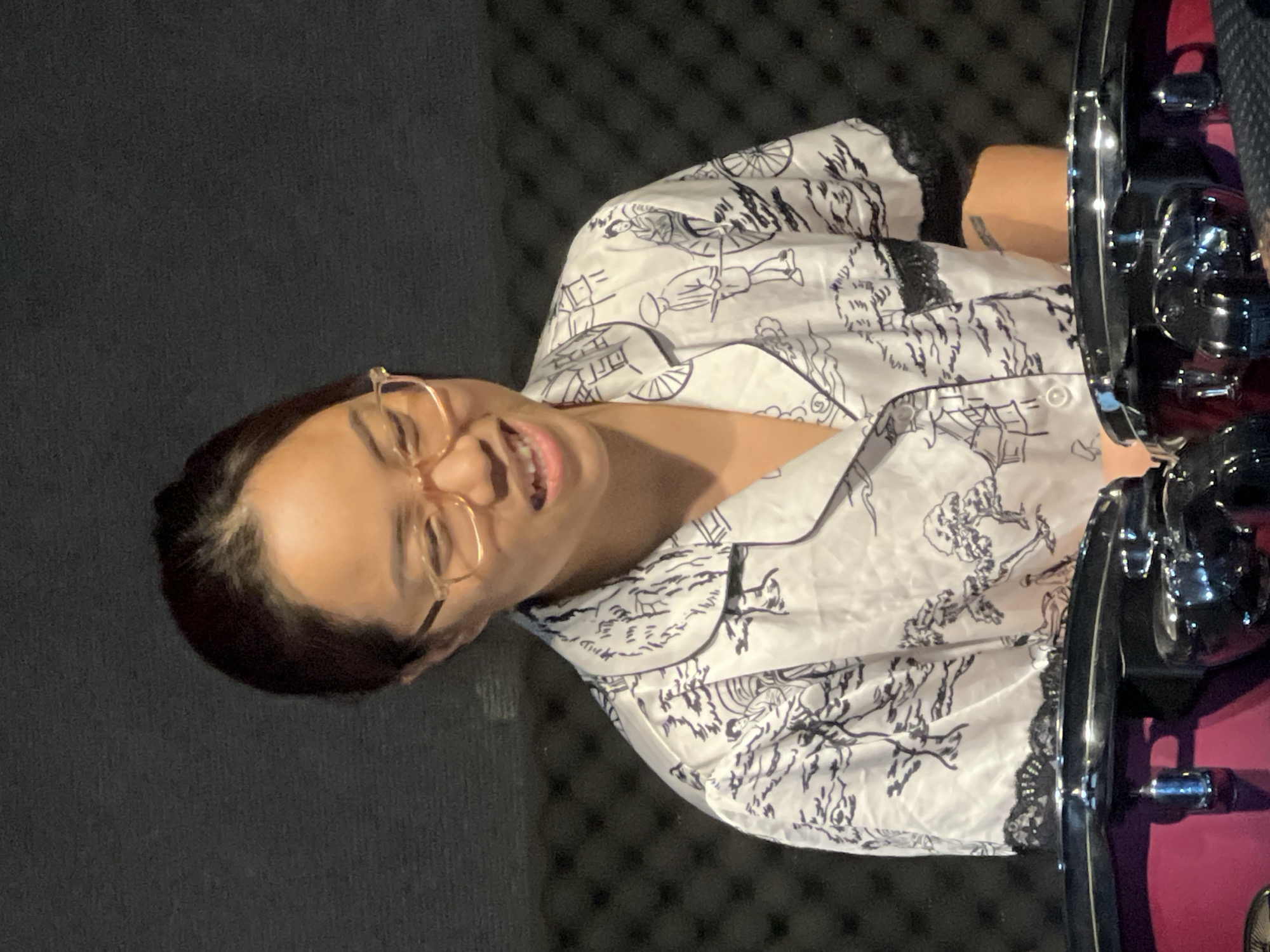 A woman with dark hair pulled back, wearing a white pajama-style shirt with a black oriental pattern and clear-rimmed glasses, smiles and looks down to her right. She is positioned in the foreground, with the upper bodies of two dark-colored drum-like instruments visible in front of her. The background consists of a textured, dark gray wall. The scene is likely indoors, possibly in a studio or practice space. No specific location in Đà Nẵng, Vietnam, can be determined from the image.
