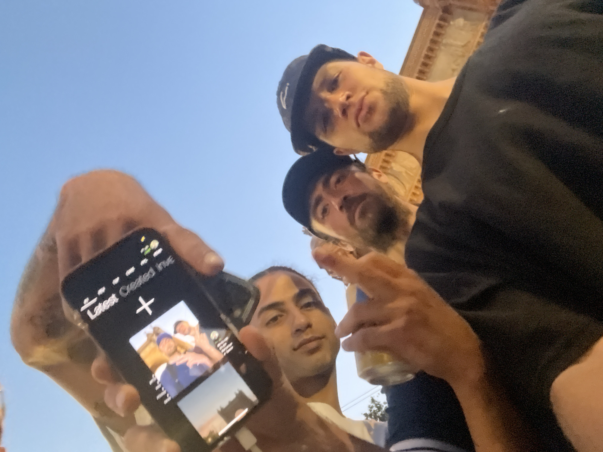 Four individuals are captured in a low-angle selfie taken outdoors, likely in a city setting. The sky above is clear and blue, suggesting daytime. In the foreground, a hand holds a smartphone, displaying a picture of two people on its screen. The phone itself has text that appears to say "Latest Created IME." Three men are visible in the shot. The man on the left, in a white shirt, is looking towards the camera. Another man, wearing a black cap, is positioned in the center, also looking towards the camera. The third man, wearing a dark baseball cap, is on the right, facing the camera with a neutral expression. One of the men is holding a can with a yellow label. The background shows a building with architectural details, hinting at an urban environment. The overall scene suggests a casual gathering or outing among friends. There are no specific visual cues to pinpoint the location in Barcelona, Spain.