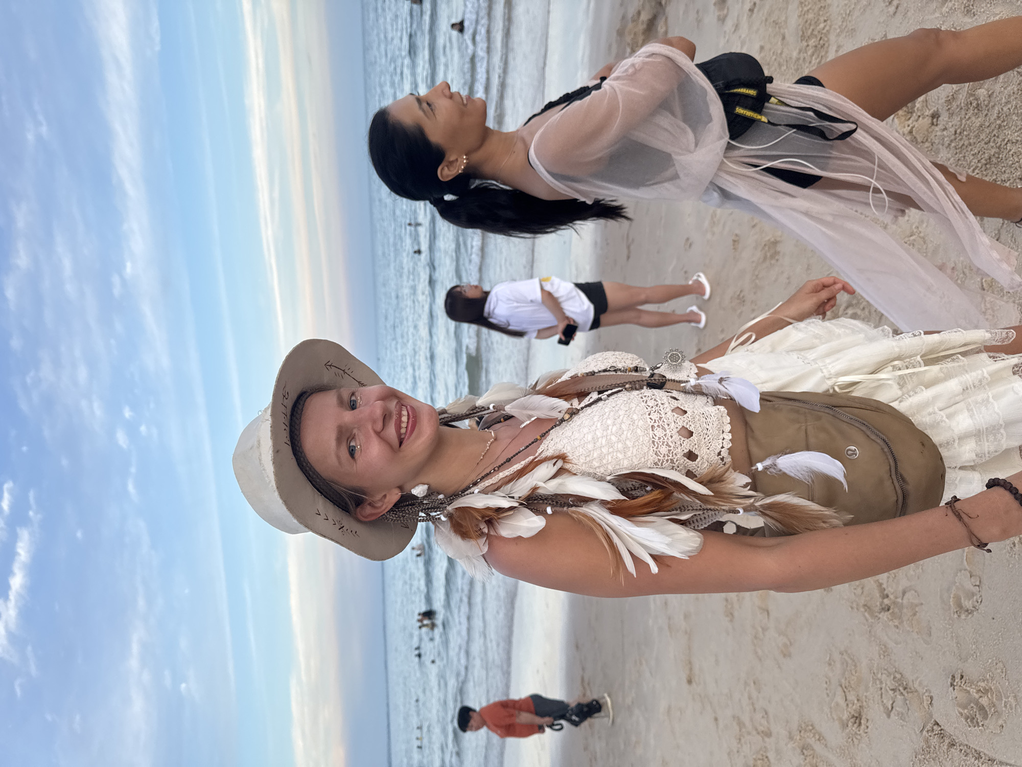 The image captures a beach scene with the ocean and sky in the background. In the foreground, a woman wearing a white, wide-brimmed hat and a white, crocheted top adorned with feathers and beads smiles at the camera. She has braided hair with feathers woven in. To her right, another woman with dark hair pulled back in a ponytail is walking away from the camera, wearing a sheer, light-colored garment over a black top and shorts. In the mid-ground, a person in an orange shirt and dark shorts is walking on the sand, and further back, another person in a white t-shirt and black shorts is also visible. Several other people are in the water. The sand on the beach appears light-colored and textured. The sky is a pale blue with wispy clouds, suggesting it is either early morning or late afternoon. The overall atmosphere appears casual and relaxed, consistent with a beach setting.
