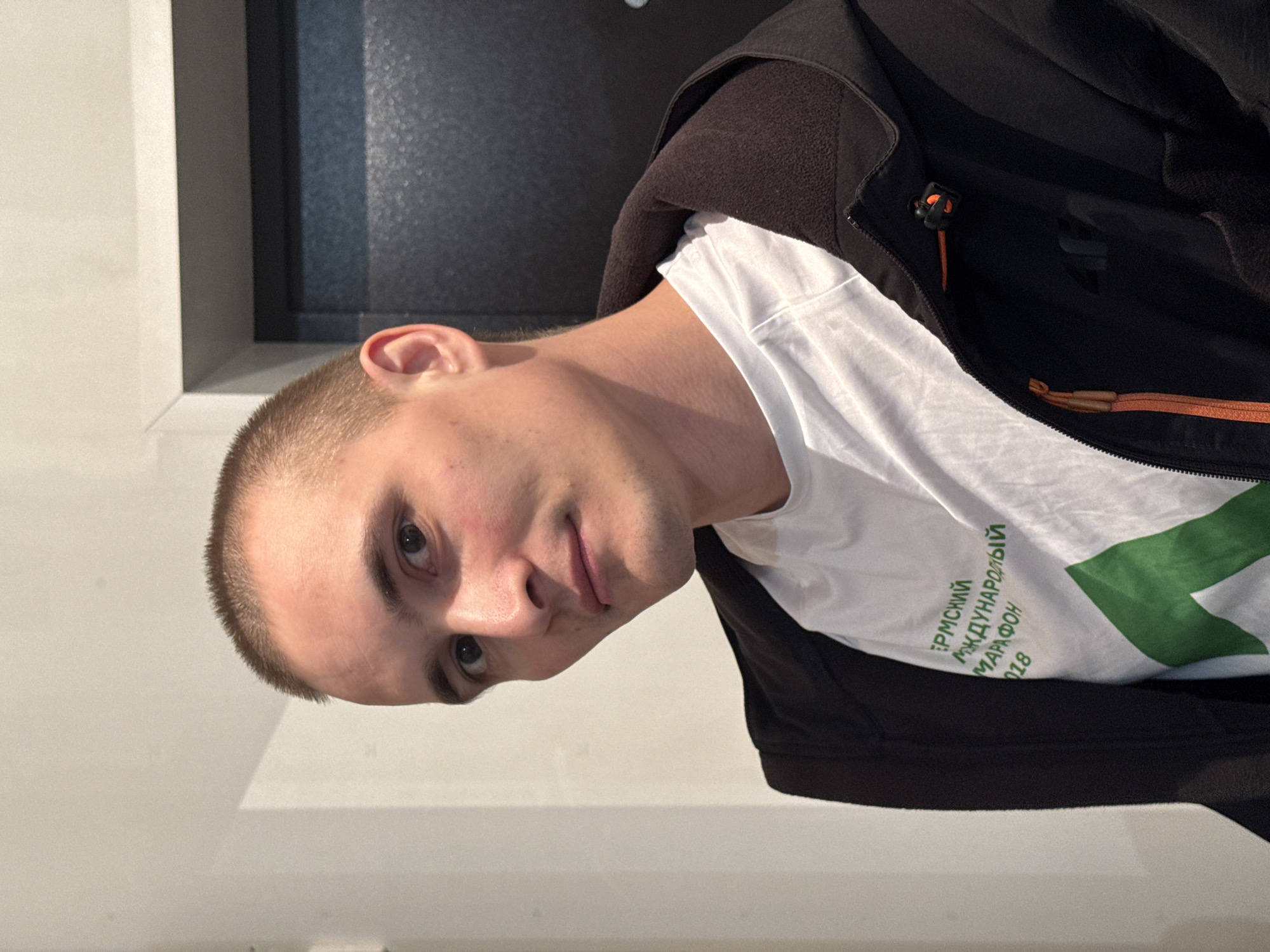 An indoor portrait captures an adult male with short, light-colored hair and dark eyes, looking directly at the camera. He wears a white t-shirt featuring green Cyrillic text, reading "ПЕРМСКИЙ МЕЖДУНАРОДНЫЙ МАРАФОН 2018" (Perm International Marathon 2018), and a green graphic. Over the t-shirt, he wears an unzipped black hooded jacket with an orange zipper pull and a dark "4F" logo on the left chest. The background consists of a light-colored wall on the left and a dark, textured door with a matching door frame on the right. The scene is located indoors; no specific external identifiers for the city of Chaikovsky, Russia, are visible.