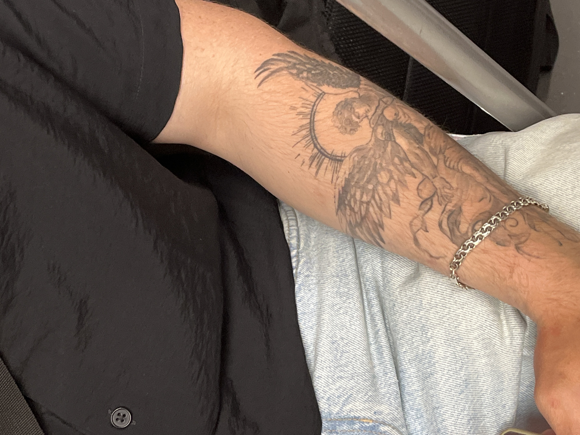 The image displays a close-up of a person's upper torso and right forearm. The individual wears a black short-sleeved shirt, visible across the chest and shoulder, with a single four-holed black button fastened near the upper left collarbone area. The shirt fabric exhibits minor creases and a slight sheen. Below the shirt, light-wash denim jeans with a noticeable diagonal weave pattern are visible, indicating a casual attire.
The right forearm is adorned with a detailed black-and-grey tattoo featuring an angelic or cherubic figure with prominent wings. A radiating sunburst pattern forms a halo-like element behind the figure's head, which is depicted looking downwards. Additional intricate tattooed designs extend further down the forearm, indicating a larger composition. A silver-colored, thick chain-link bracelet is worn on the wrist.
In the background, a dark, possibly fabric-covered surface is partially visible to the right, adjacent to a smooth, light grey, metallic bar, which could be part of a seat or railing. The lighting is uniform, suggesting an indoor or shaded environment. The specific location in the city of Minsk, Belarus, cannot be ascertained from the visual evidence in the image.