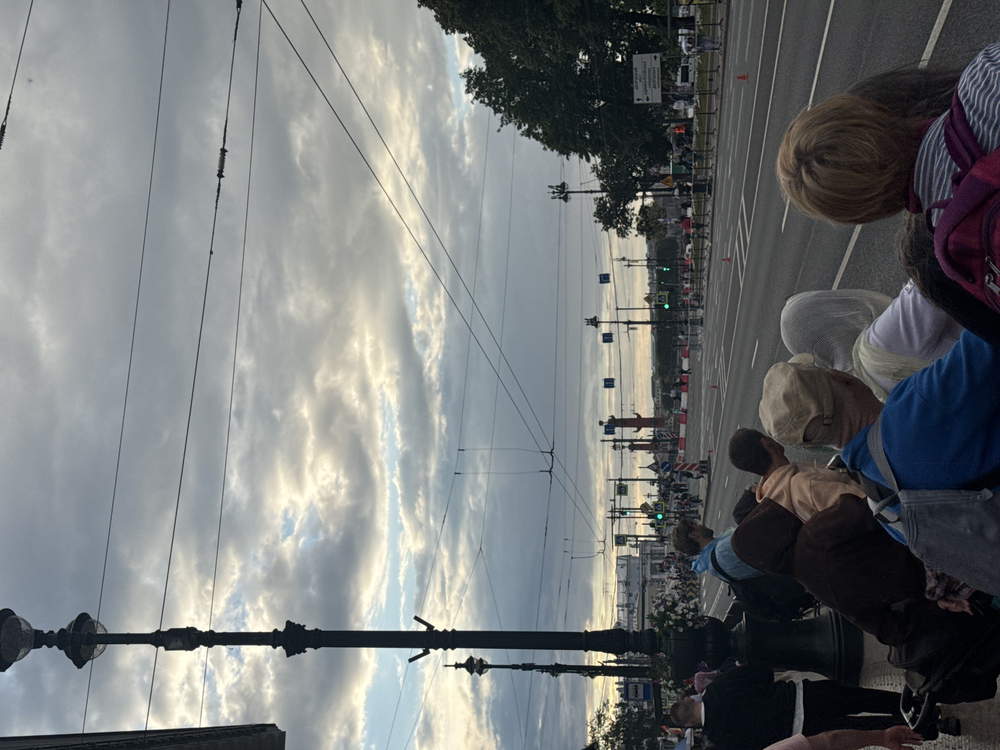 This image captures a view of Palace Square in Saint Petersburg, Russia, under a cloudy sky. In the foreground, a line of people are waiting, some with luggage. They are positioned on a sidewalk next to a street where traffic is moving and people are walking. Prominent in the scene are several large, ornate black lampposts. In the background, the distinctive Rostral Columns are visible, along with other architectural elements of the square. The overall atmosphere suggests a public gathering or event is taking place.