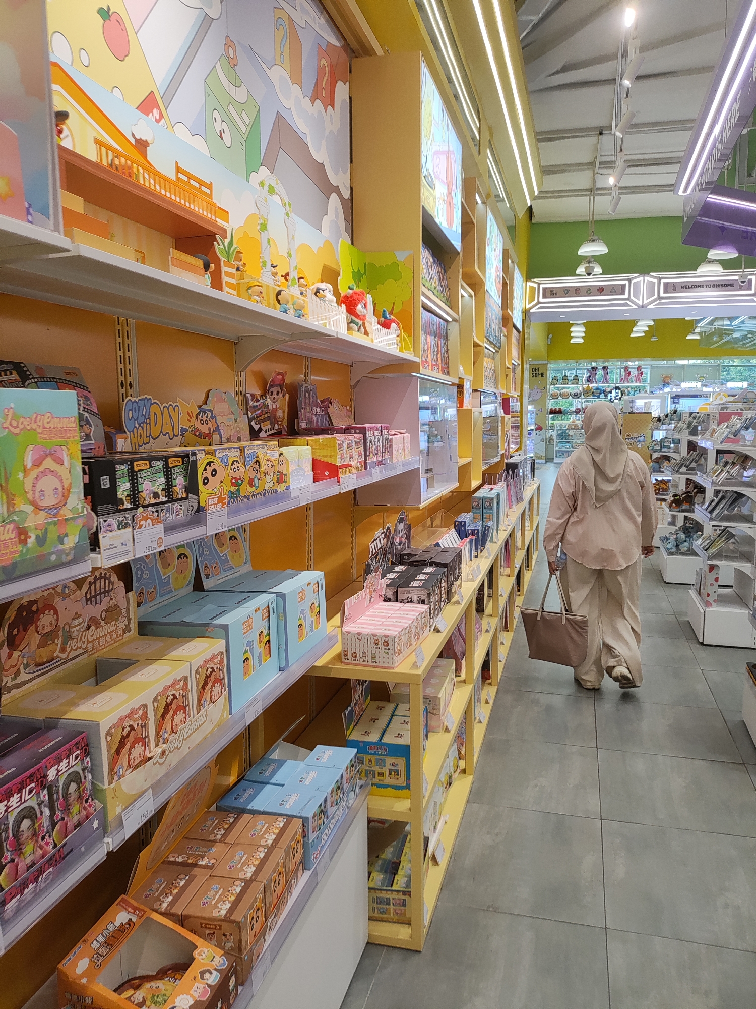 This image shows the interior of a brightly lit store, likely a toy or novelty shop, filled with colorful merchandise. The shelves are stacked high with various boxed items, many featuring cartoon characters and playful designs. In the foreground, a person wearing a light-colored hijab and loose-fitting clothing is walking away from the camera, carrying a large tote bag. They are walking down an aisle lined with shelves on both sides. The store environment appears clean and organized, with modern lighting fixtures visible on the ceiling. The overall impression is of a lively and engaging retail space. Based on the provided context that this image is from Lambangjaya, Indonesia, this scene depicts a retail establishment within that city.