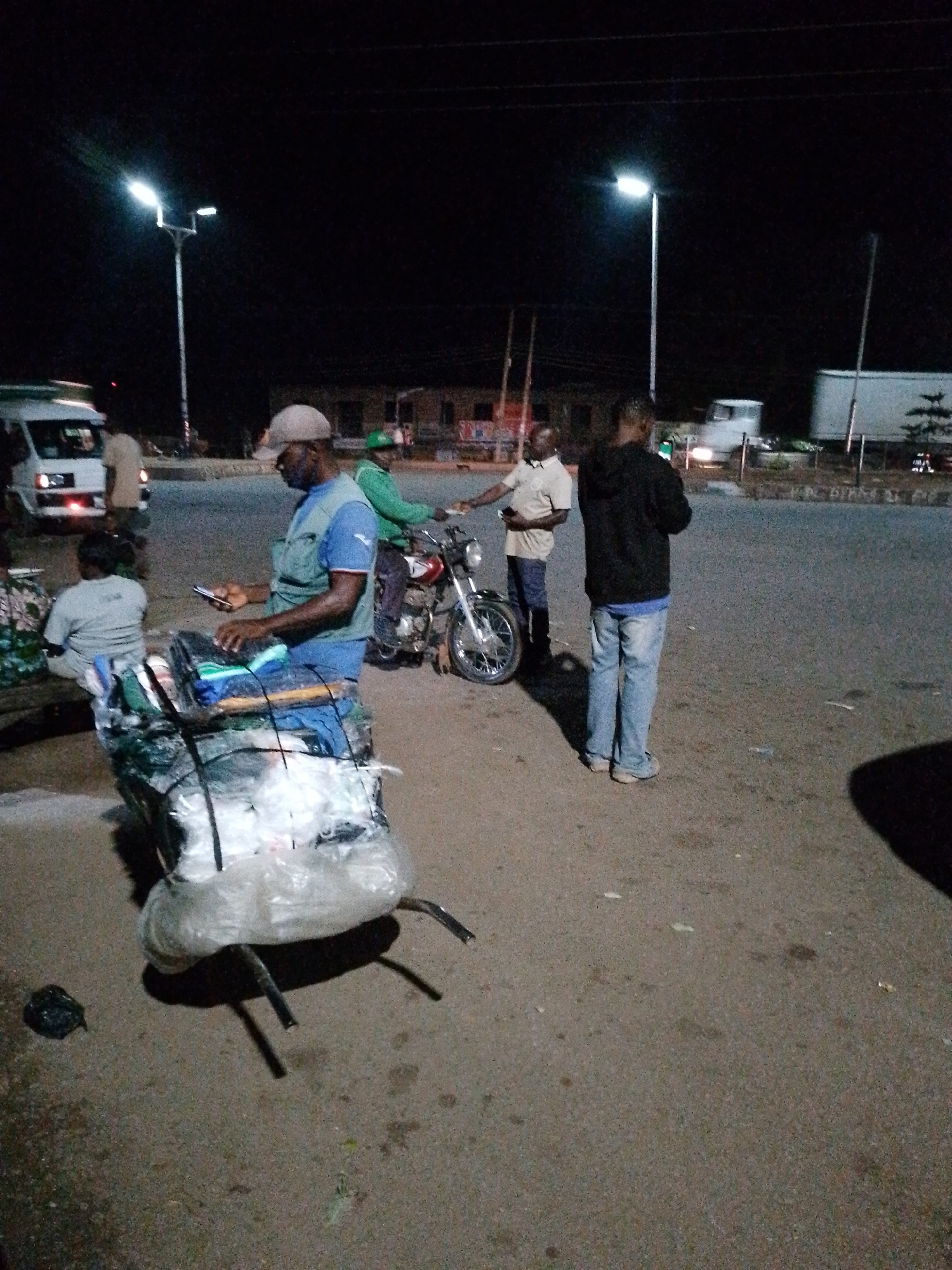 At night, a group of people are gathered on a street in an urban environment. In the foreground, a man wearing a blue vest and a cap is looking at his phone while standing next to a cart piled high with goods wrapped in plastic. The cart is supported by two metal legs. To his right, a motorcycle is parked. Behind the motorcycle, two men are conversing, one in a light-colored shirt and the other in a black hoodie. Further in the background, a white van is visible, and a street lamp illuminates the scene. Buildings and other vehicles are also present in the background. The ground is paved asphalt.