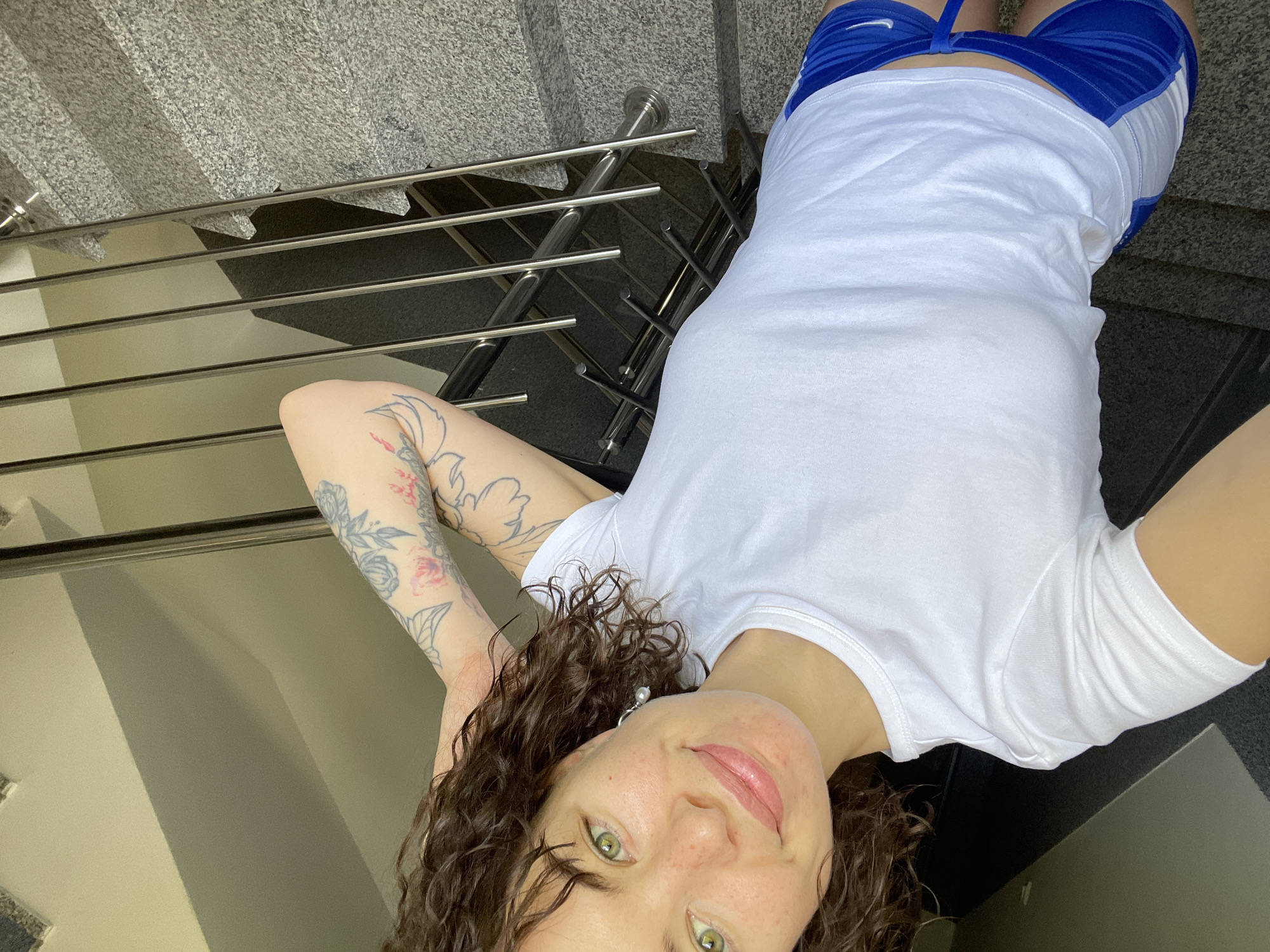 A young woman with dark, curly hair, green eyes, and freckles is the central subject of the image. She has a slight, relaxed smile and is looking directly at the camera. She wears a plain white short-sleeved t-shirt and blue athletic shorts with white panels, featuring a visible Nike swoosh logo on the left thigh. Floral tattoos, with some pink coloring, are visible on both of her arms. A white pearl drop earring is seen on her right ear.
The woman is positioned as if lying down or leaning back on a surface, with her arms raised and bent, possibly resting behind her head. The photo is taken from a low angle, looking upwards at her.
The setting appears to be an indoor or covered area, likely a stairwell, in Boreti, Montenegro. Behind her are stairs made of a light-grey, speckled stone, resembling granite. A modern metal railing with multiple horizontal bars runs alongside the staircase. Parts of a light-colored wall and darker areas, possibly shadows or reflections from the railing structure, are also visible in the background. The lighting is soft and even, suggesting a well-lit interior during the daytime.